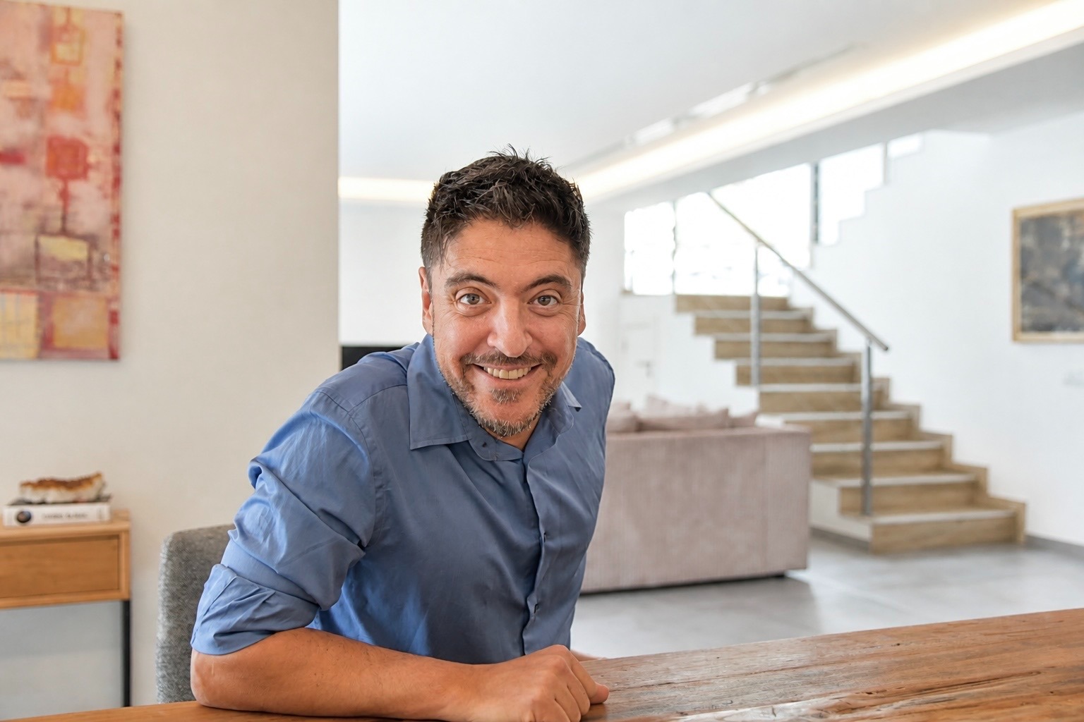 Smiling man in a blue shirt sitting at a wooden table in a bright modern home office.