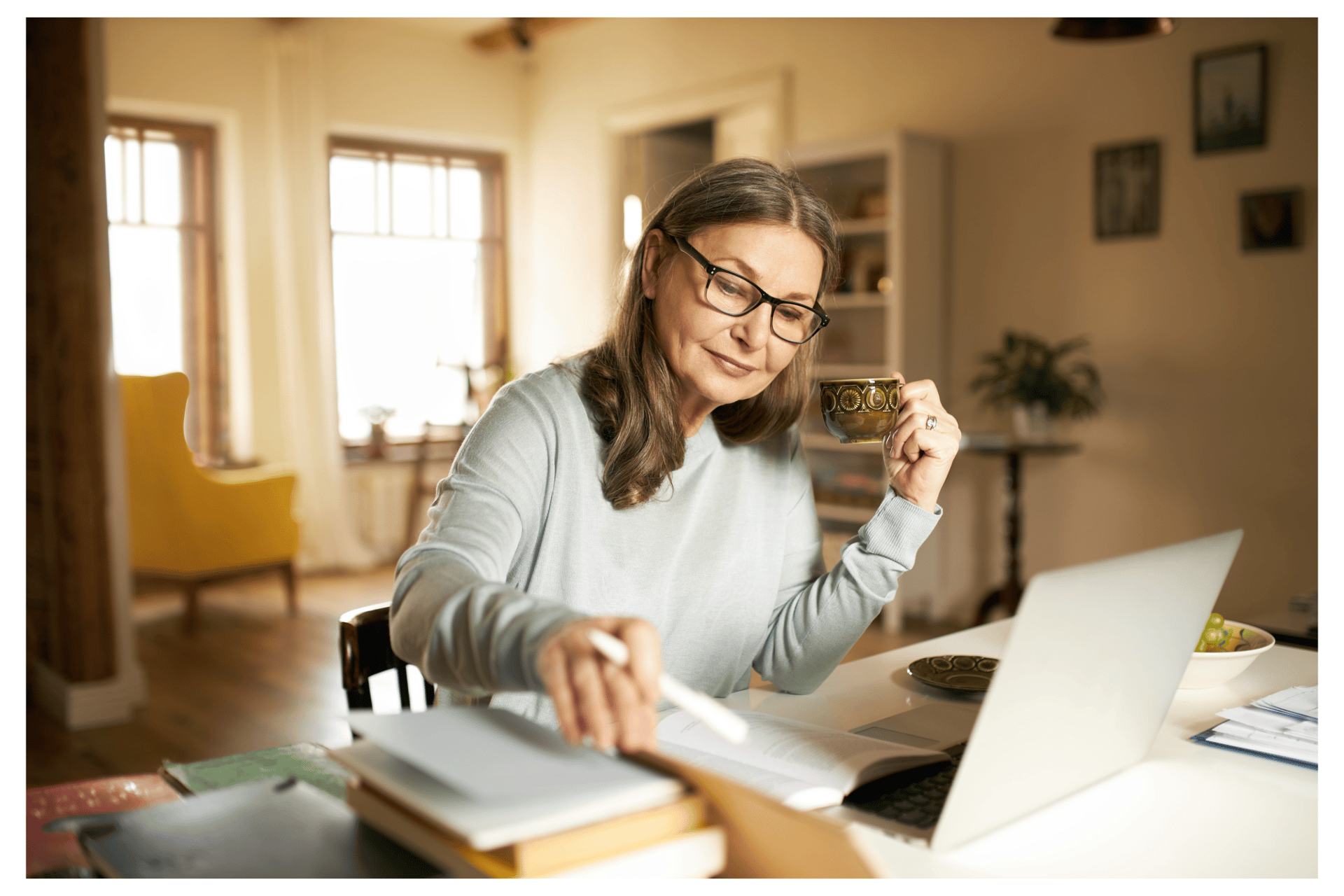 A senior woman wearing glasses drinks tea while working on a laptop at her home office desk.