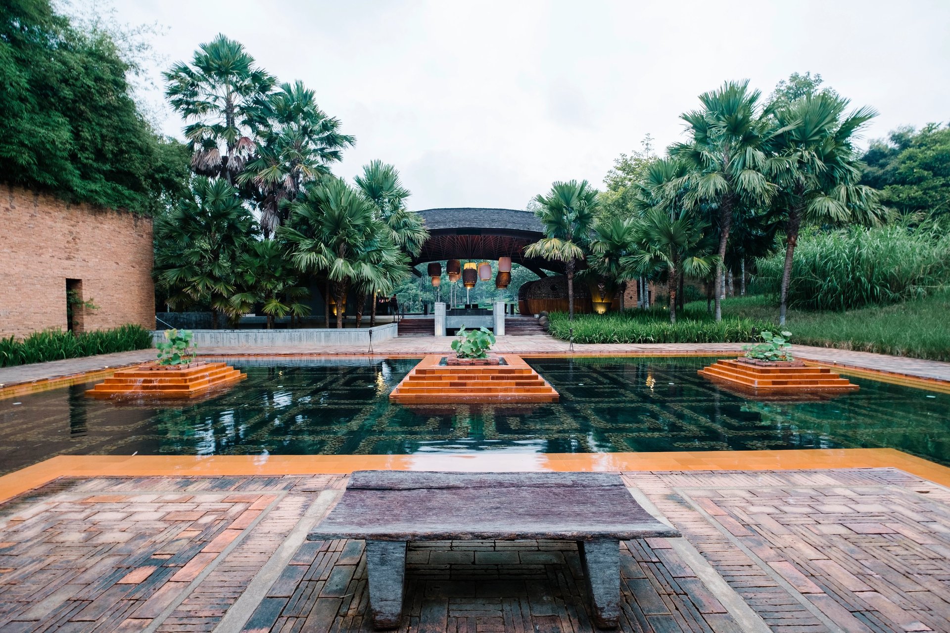 Luxury tropical resort courtyard featuring a tiered reflection pool, palm trees, and stone architecture.