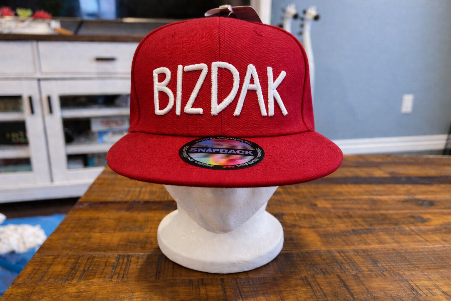 A red snapback hat with white embroidered BIZDAK text displayed on a foam head over a wooden table.