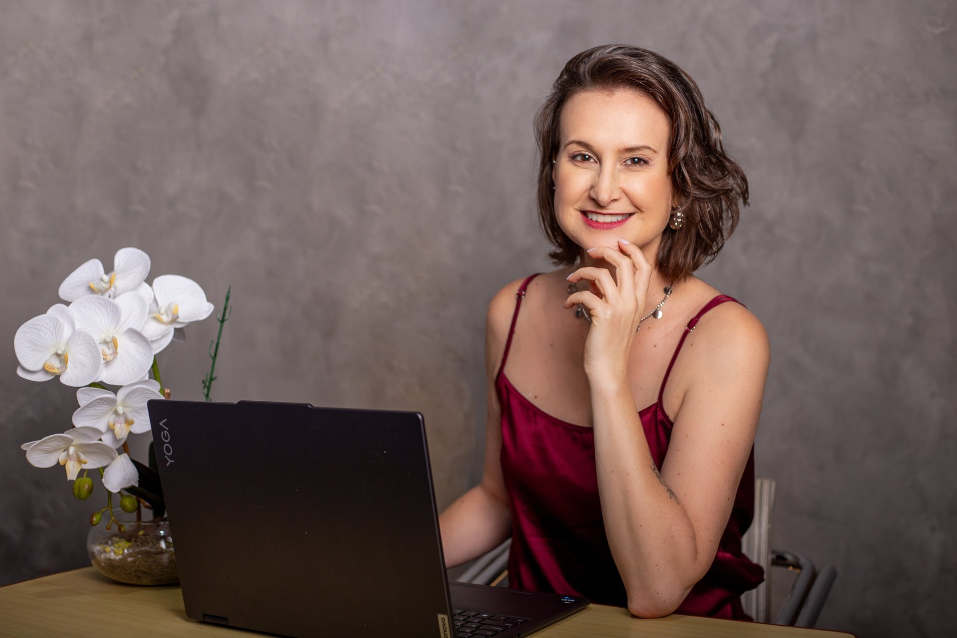 Mulher sorrindo, de cabelo castanho curto, sentada em uma mesa com a mão no queixo.