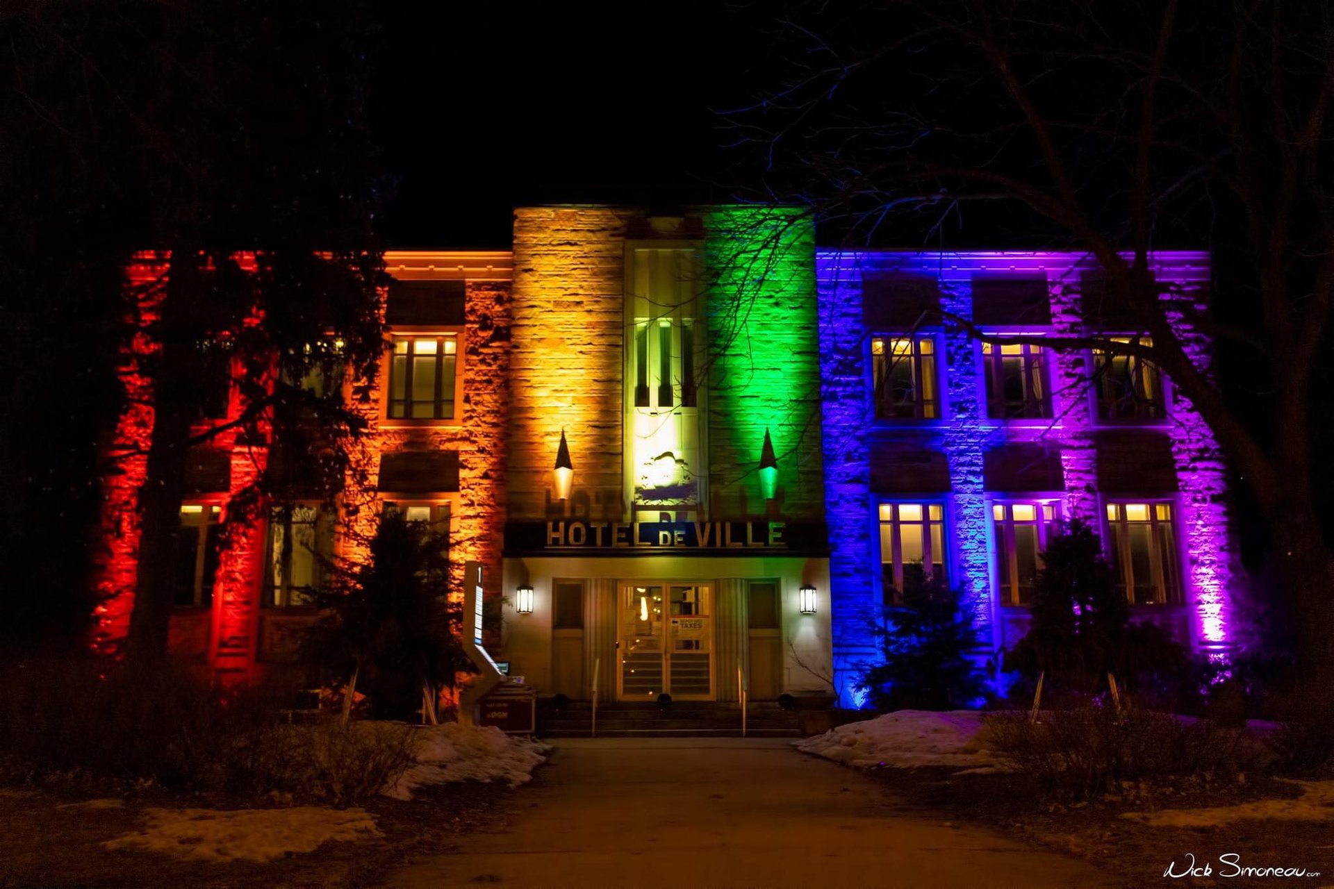 The Hotel de Ville building illuminated with vibrant rainbow pride colors at night.