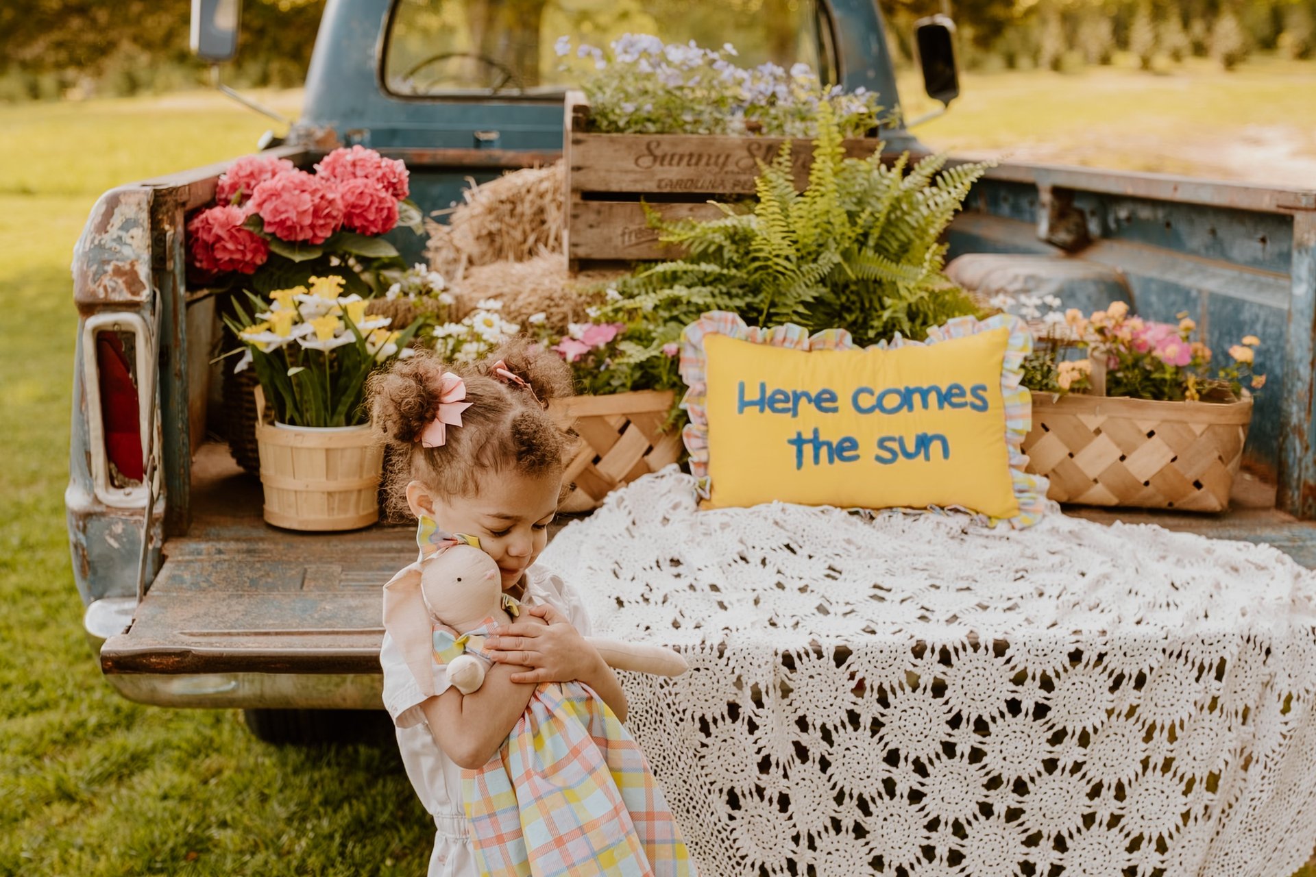 A toddler holding a doll stands near a vintage blue truck bed filled with spring flowers and a yellow sun pillow.