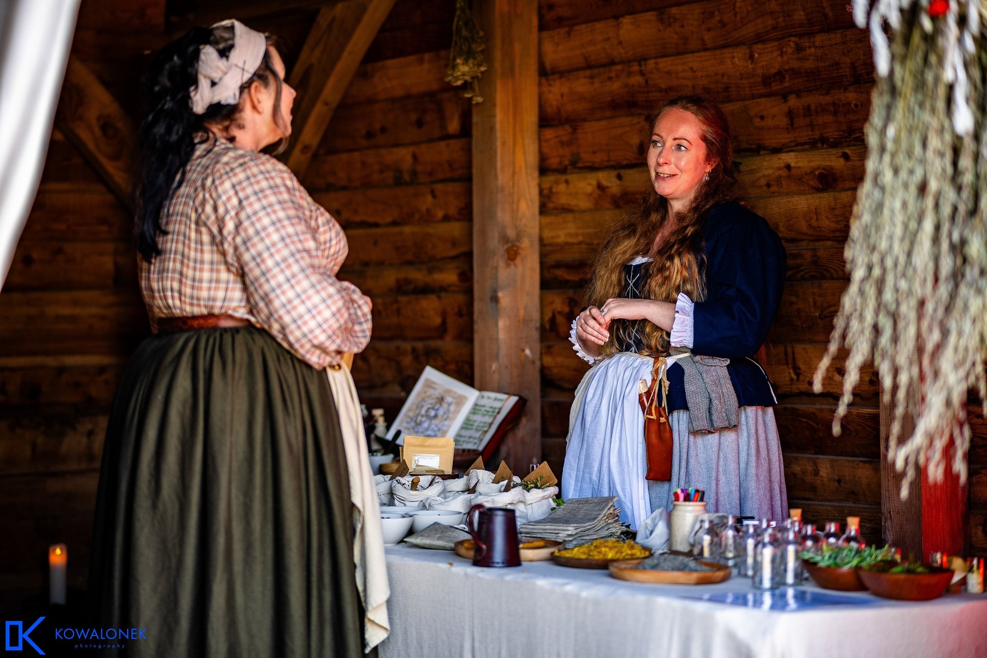 a woman in 18th century clothing stands behind a table covered in herbal remedies