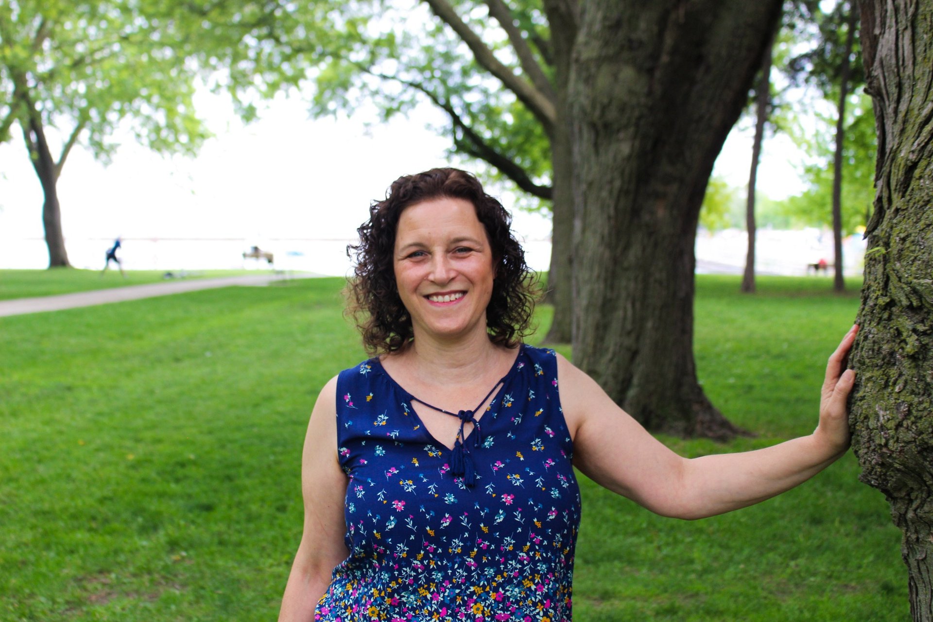 Picture of Natanya in a park, hand on a tree, water in the background.
