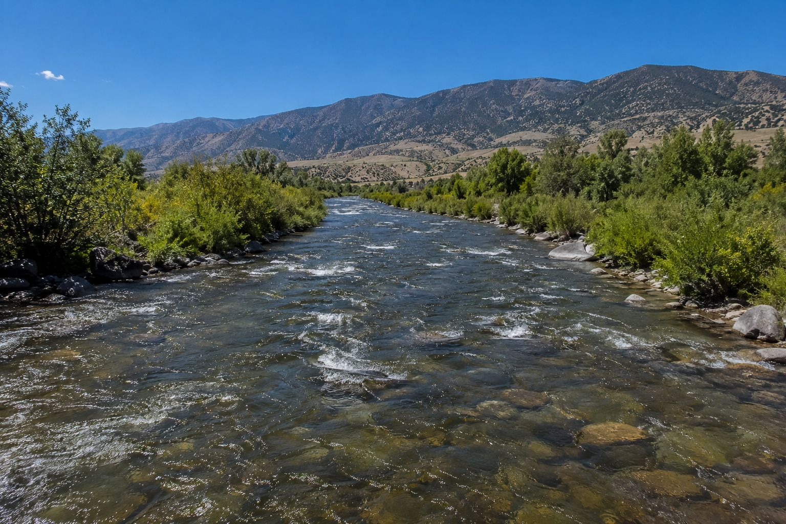 A wide river with clear water flowing through a green valley with mountains under a blue sky.