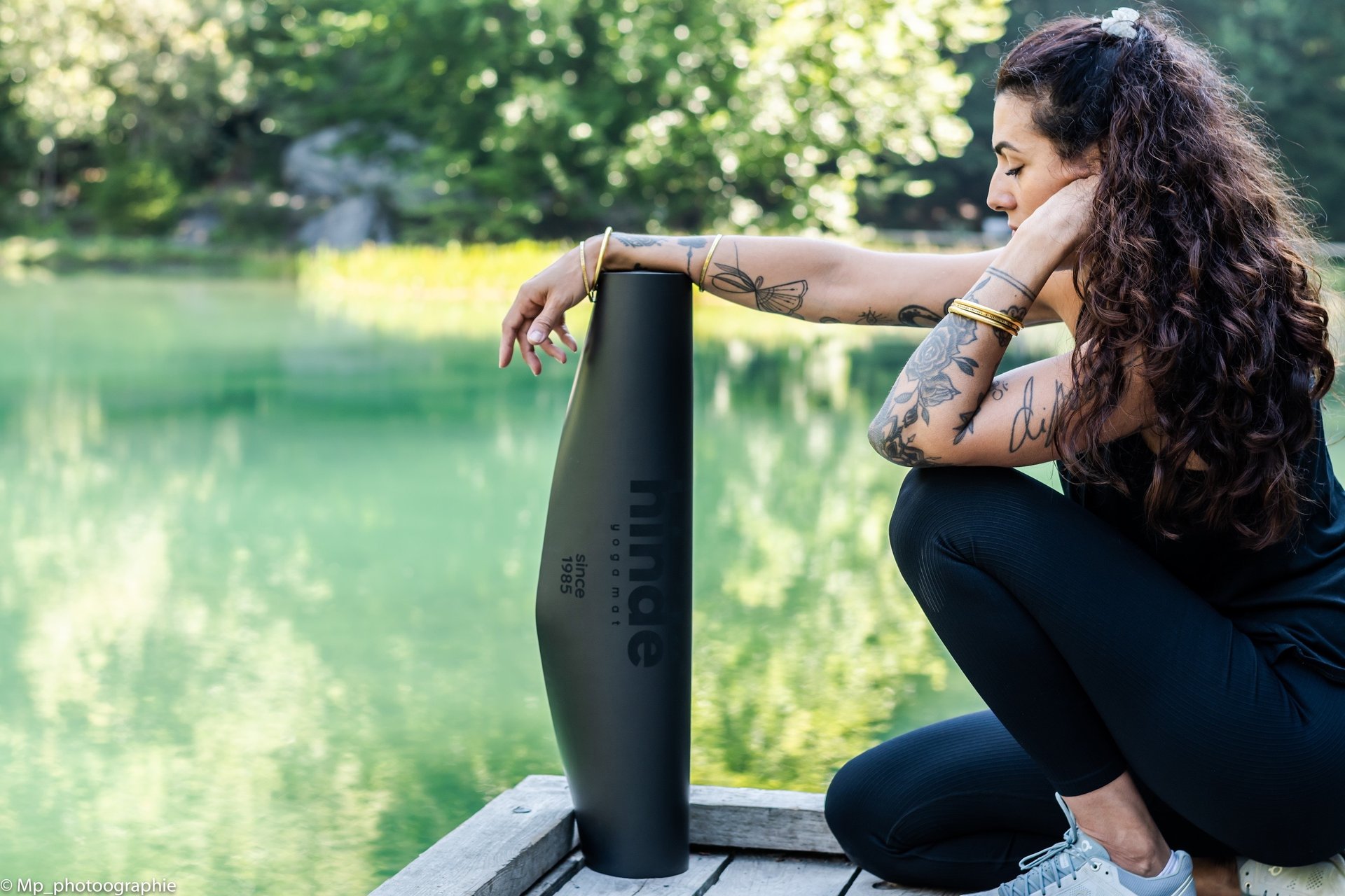 Tattooed woman posing with a black eco-friendly yoga mat on a wooden dock by a green lake.