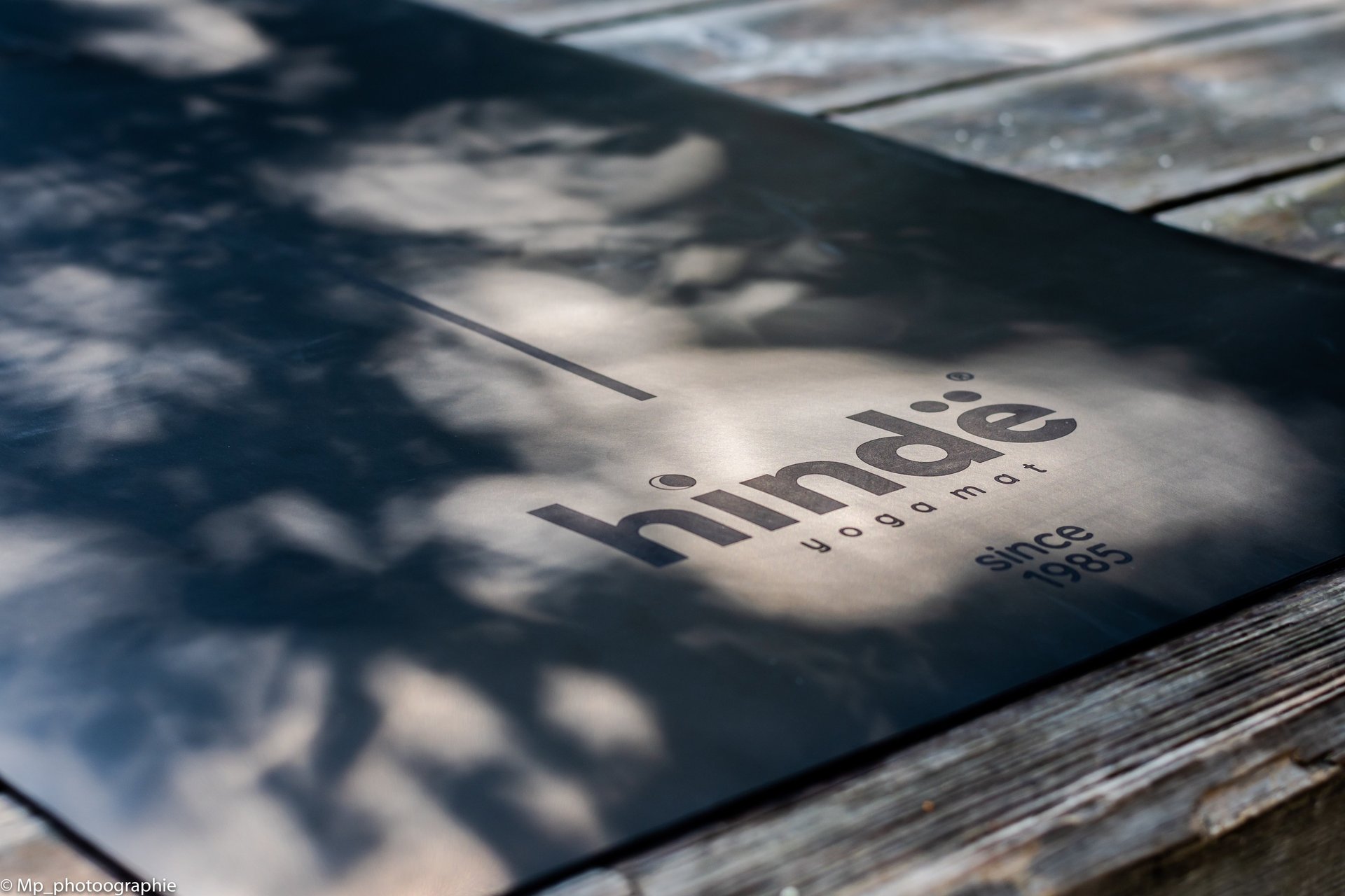 Black Hindie yoga mat with alignment lines on a rustic wooden deck with natural leaf shadows.