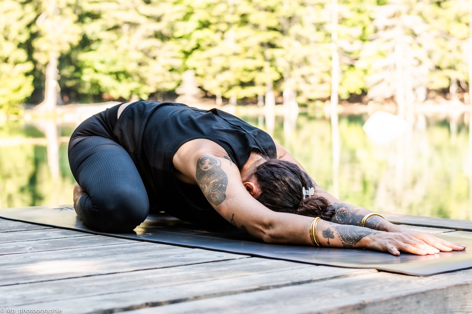 Tattooed woman practicing child's pose yoga on a wooden dock by a calm lake in the forest.