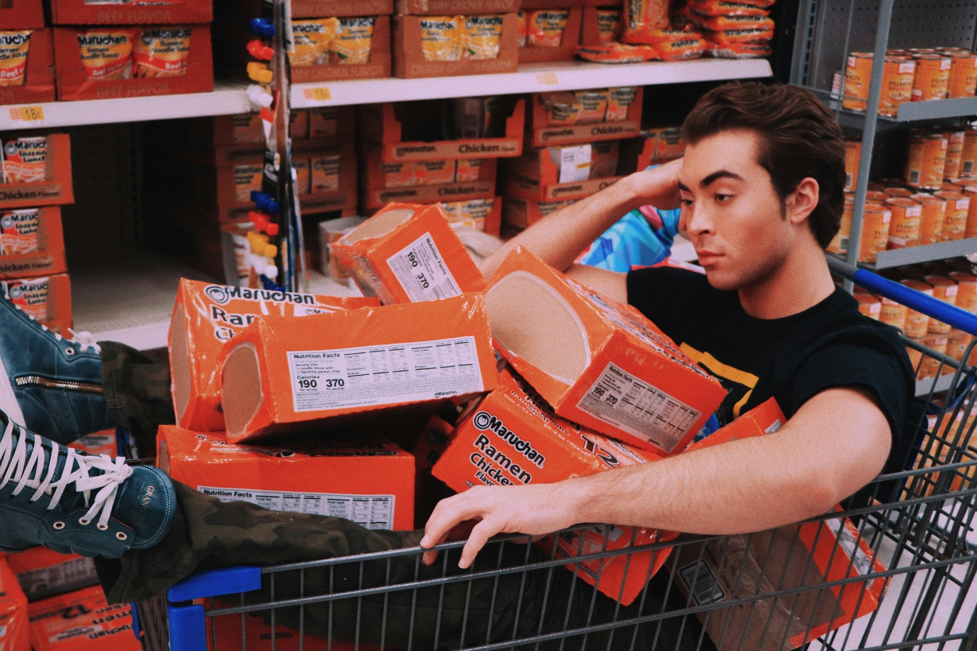 A man sits in a shopping cart filled with orange Maruchan chicken ramen noodle soup boxes in a grocery store aisle.