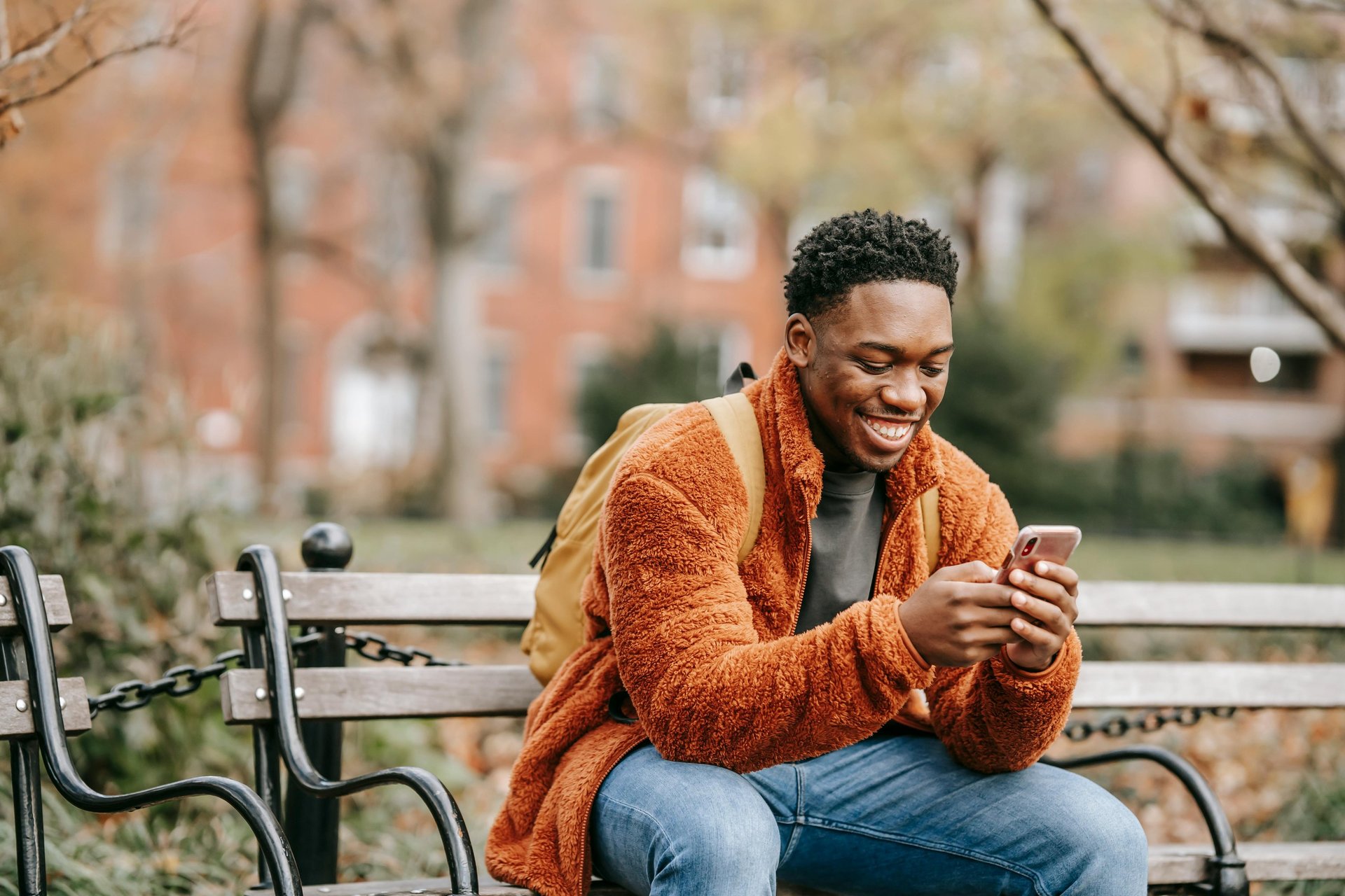 A smiling young man in a fleece jacket using a smartphone while sitting on a park bench.