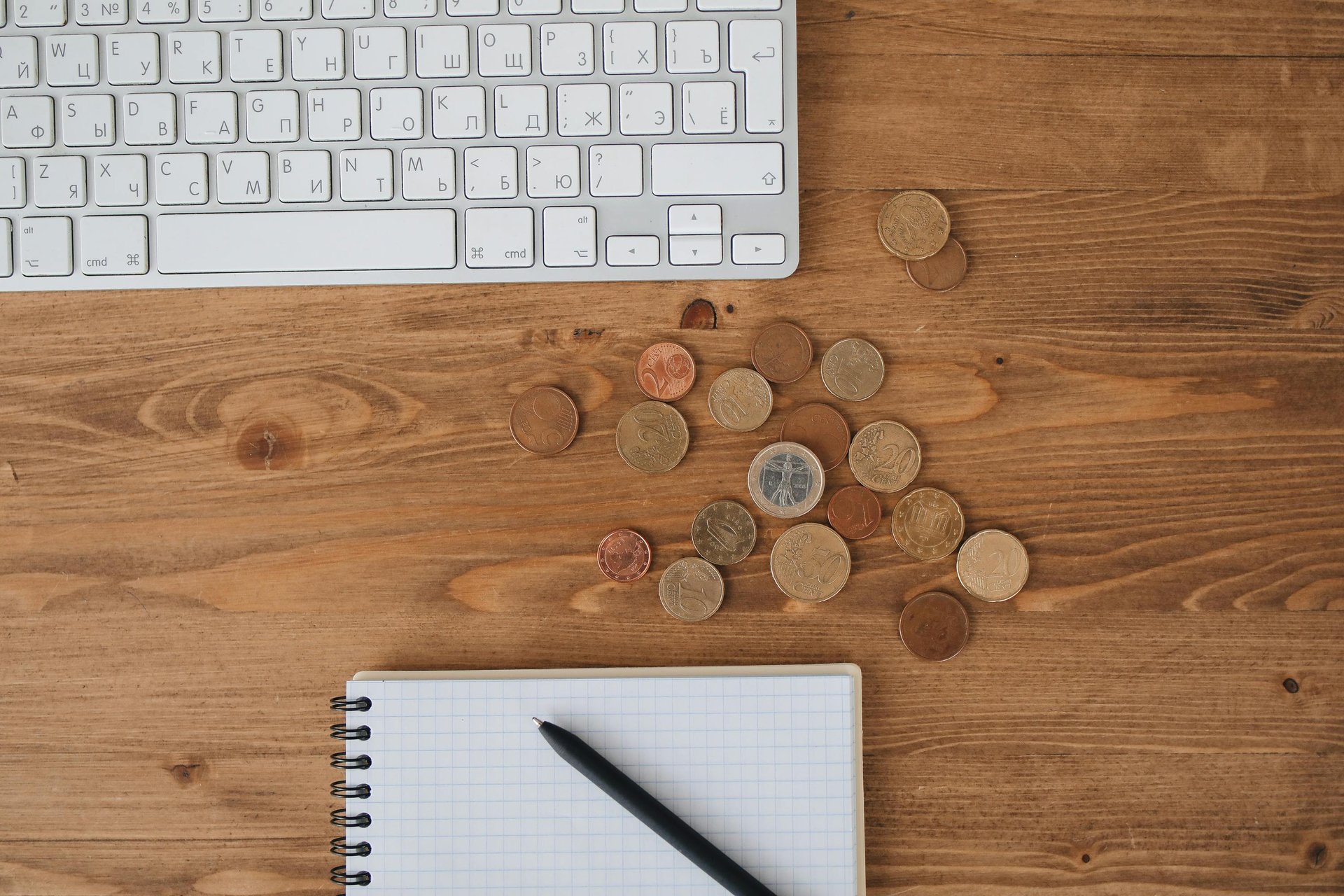 Overhead view of a computer keyboard, Euro coins, and a notebook with a pen on a wooden desk.