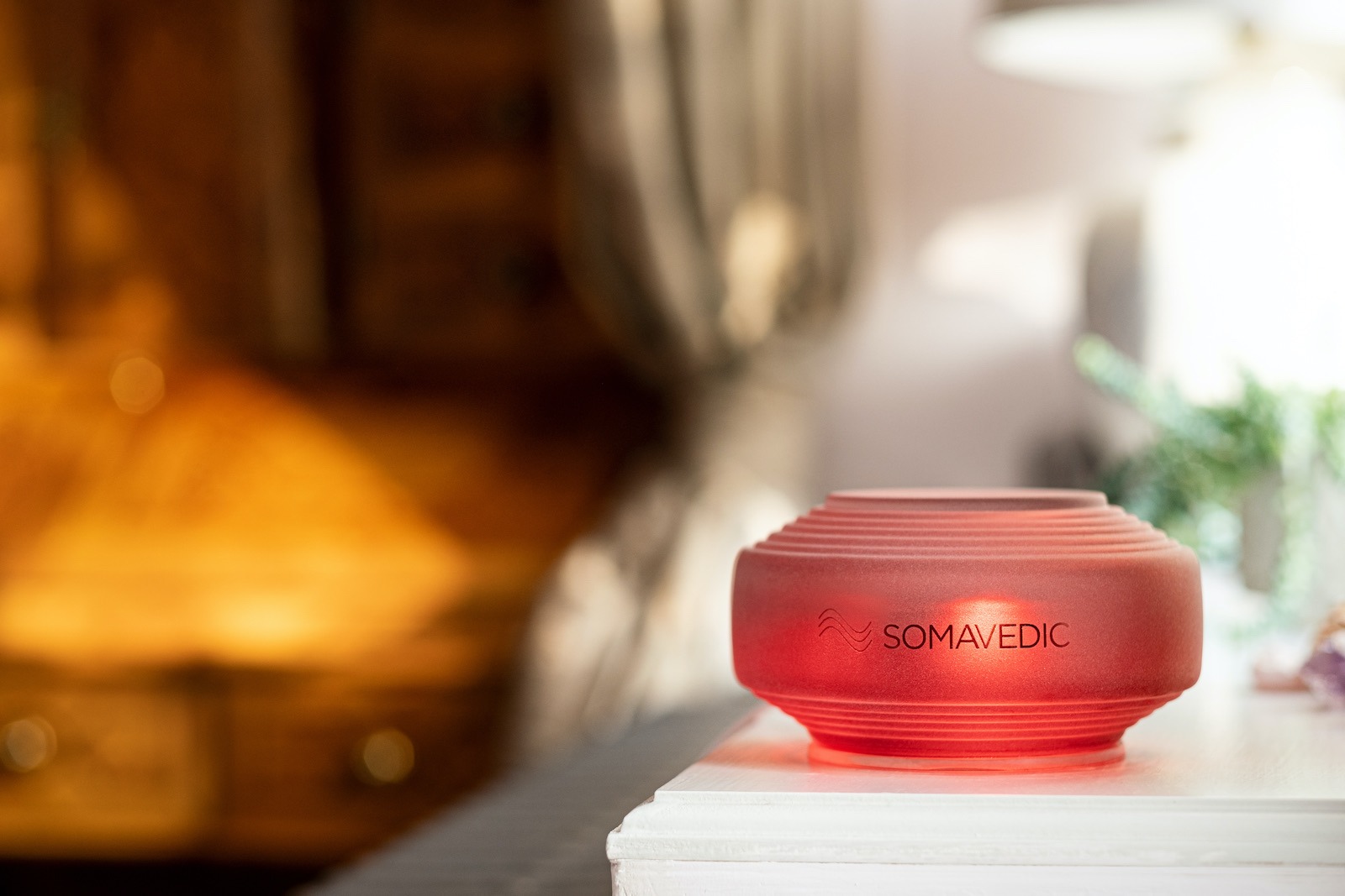 A glowing red Somavedic EMF mitigation device on a white nightstand in a cozy bedroom.