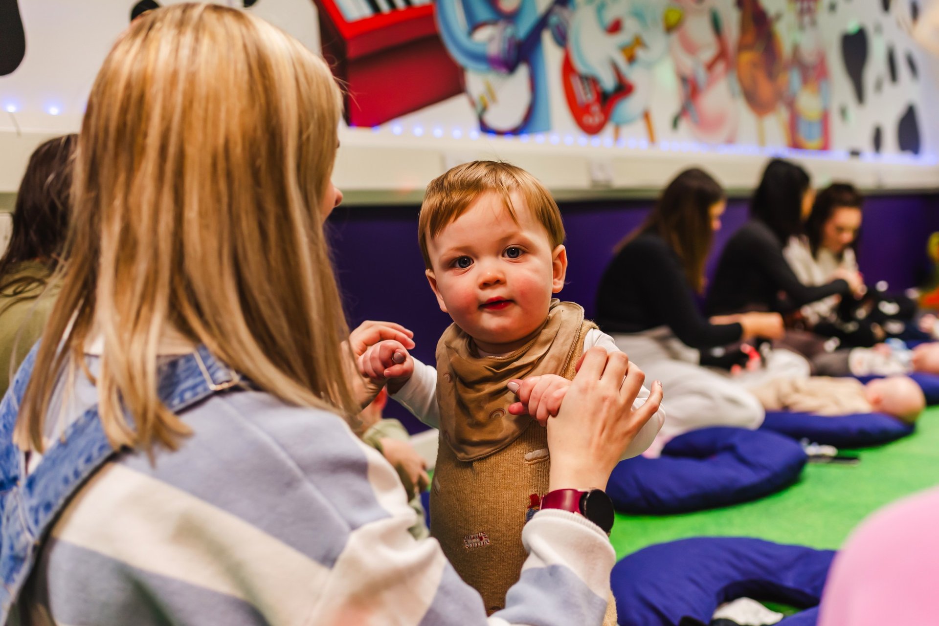 A young boy staring into the camera, holding his mums hands to stand up in moo class.