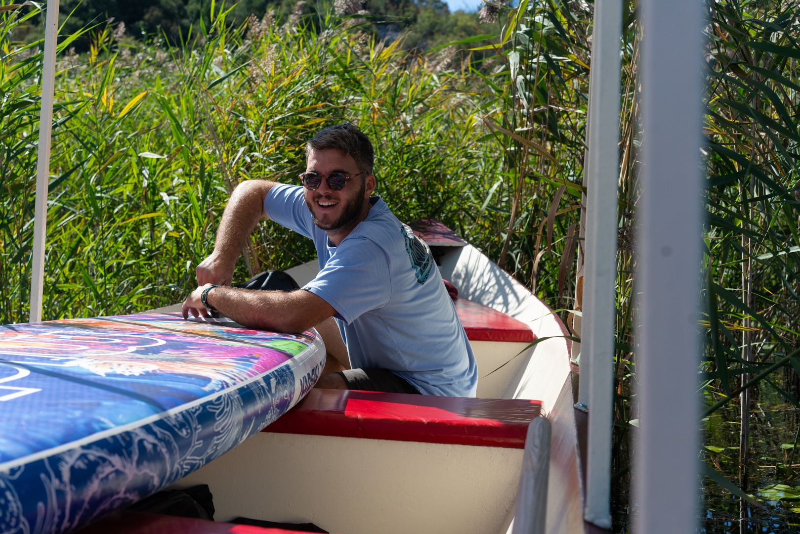 A smiling man with sunglasses sits in a boat with a colorful paddleboard near tall reeds.