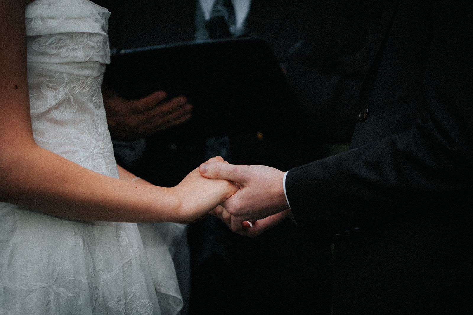 Close-up of a bride in a lace gown and groom in a black suit holding hands during a wedding ceremony.