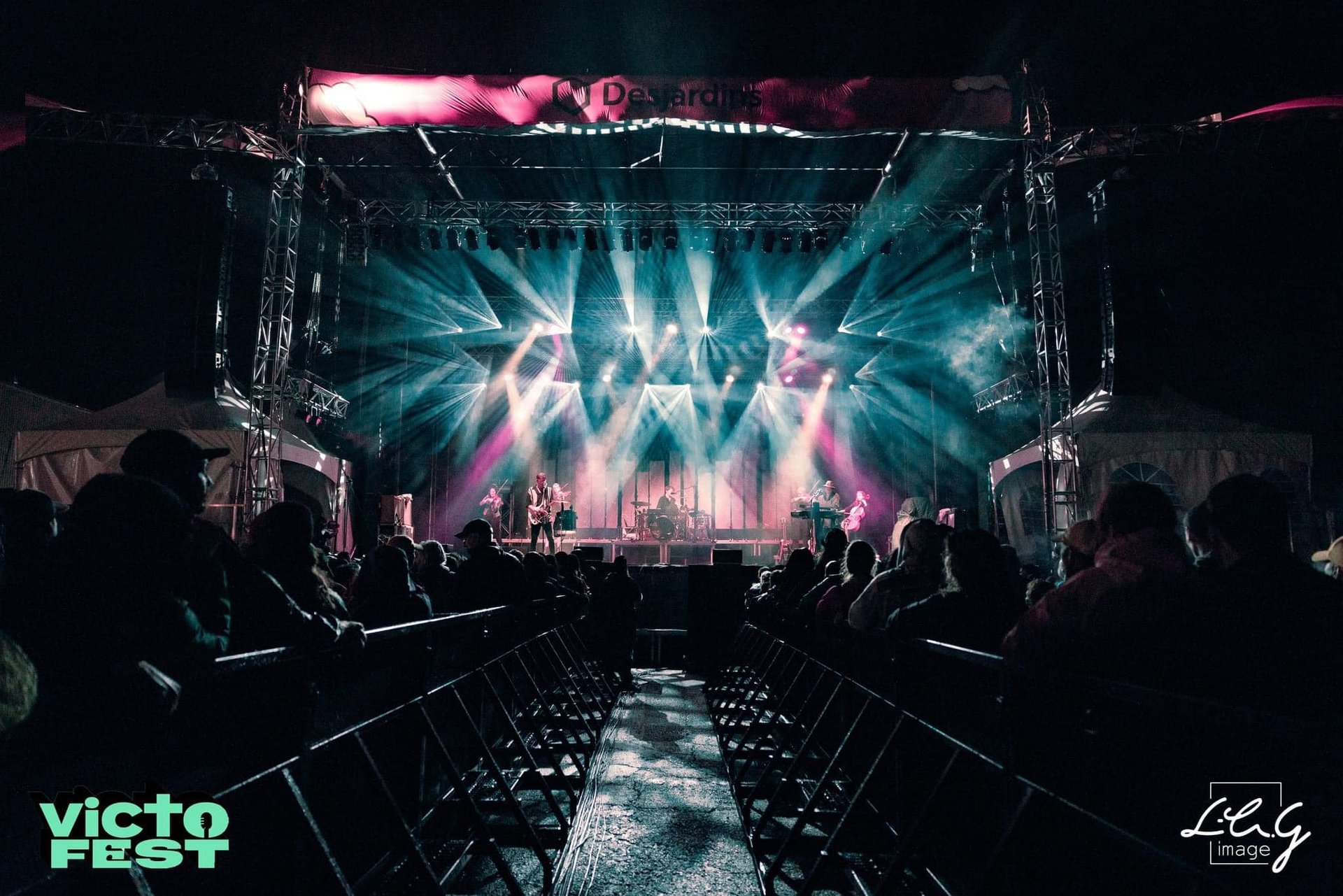 A band performs on a brightly lit outdoor stage with dramatic spotlights at VictoFest music festival.