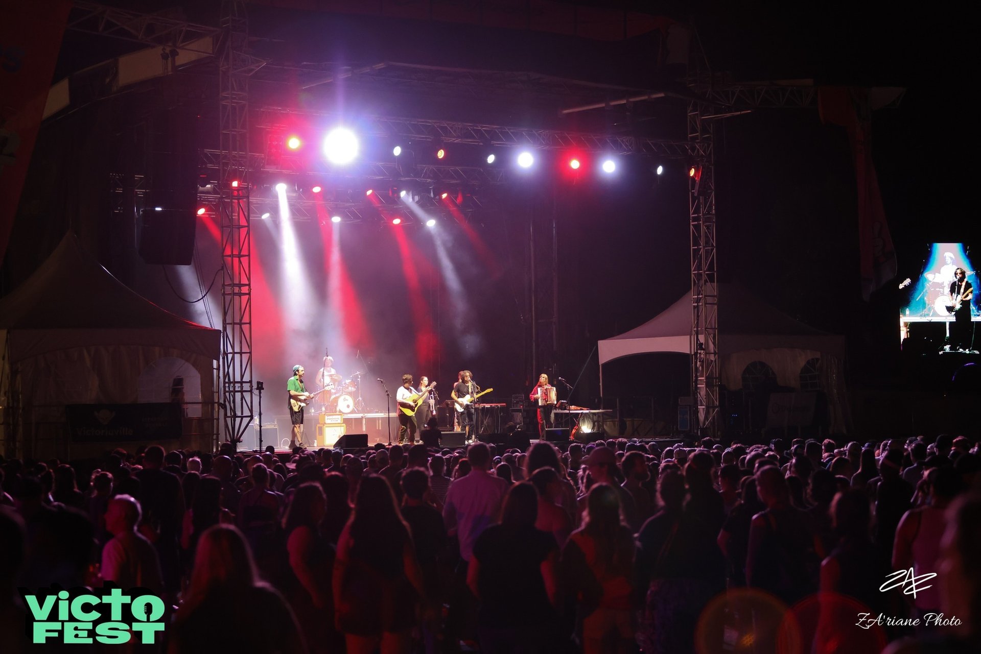 A live band performs on a brightly lit outdoor stage for a large crowd at VictoFest music festival.
