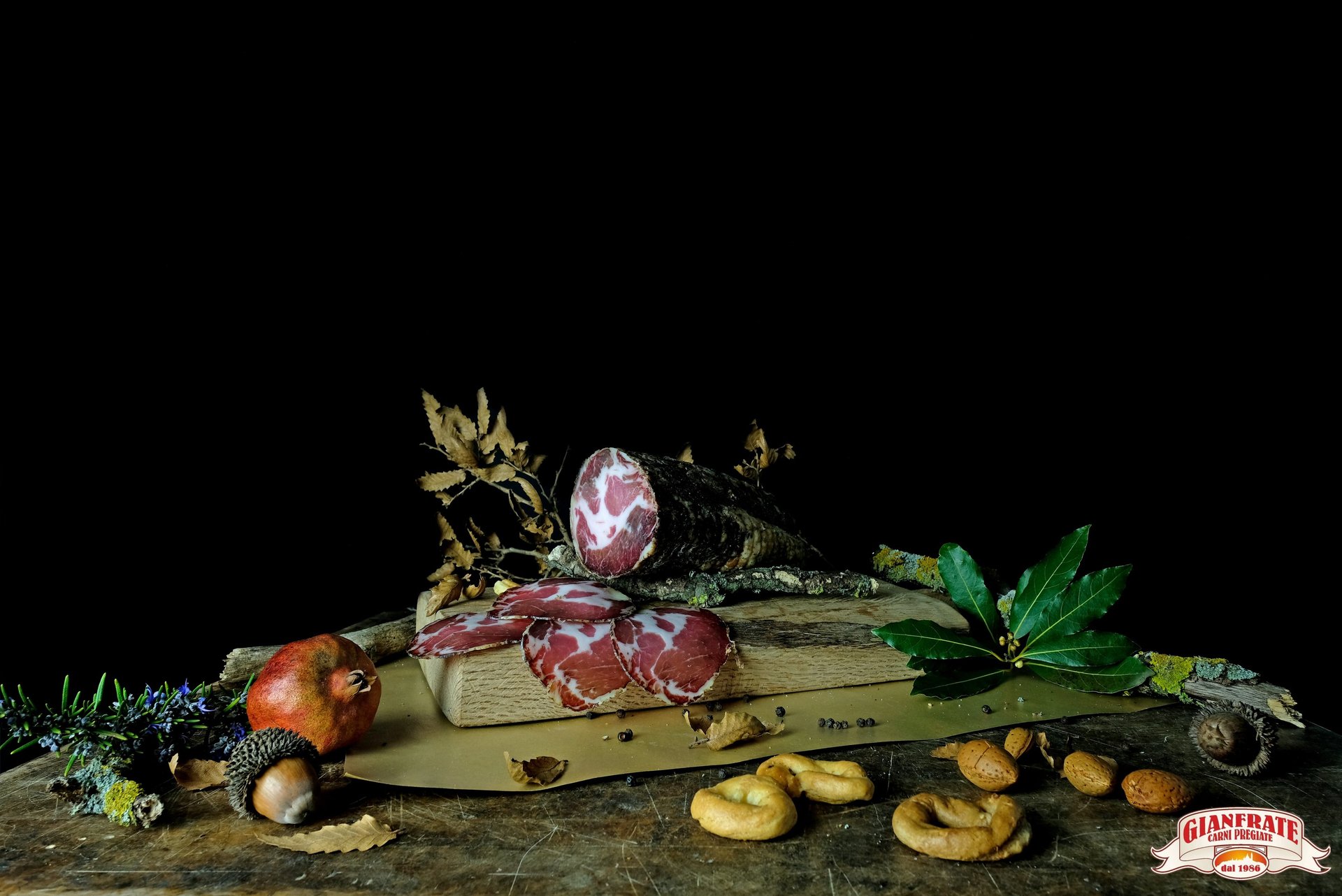 Slices of cured capocollo ham on a rustic wooden board with taralli, almonds, and pomegranate.
