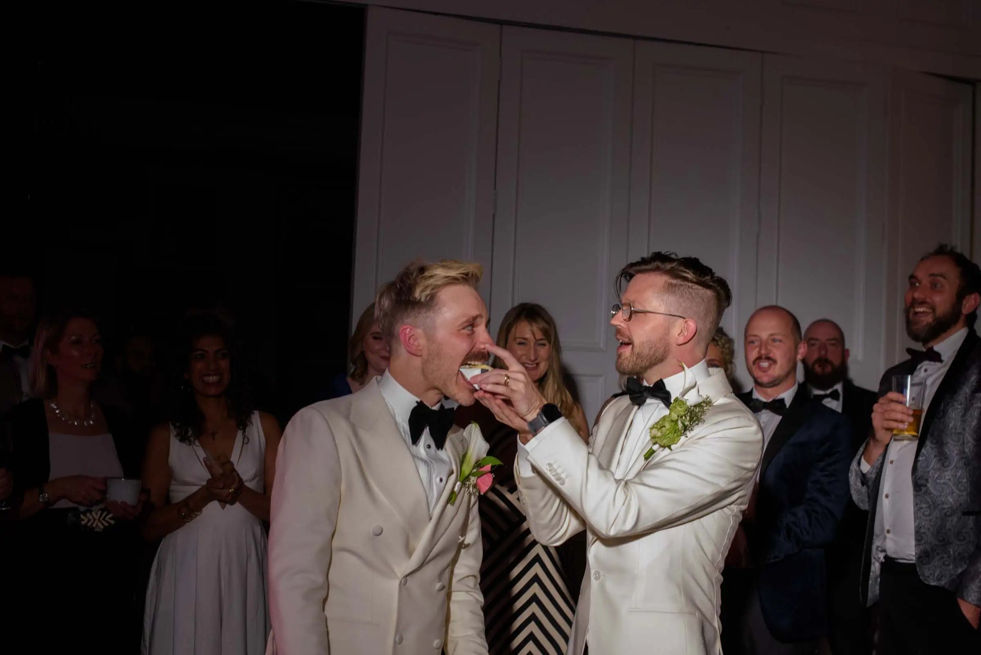 Groom in a white tuxedo feeding cake to his husband at their formal wedding reception.
