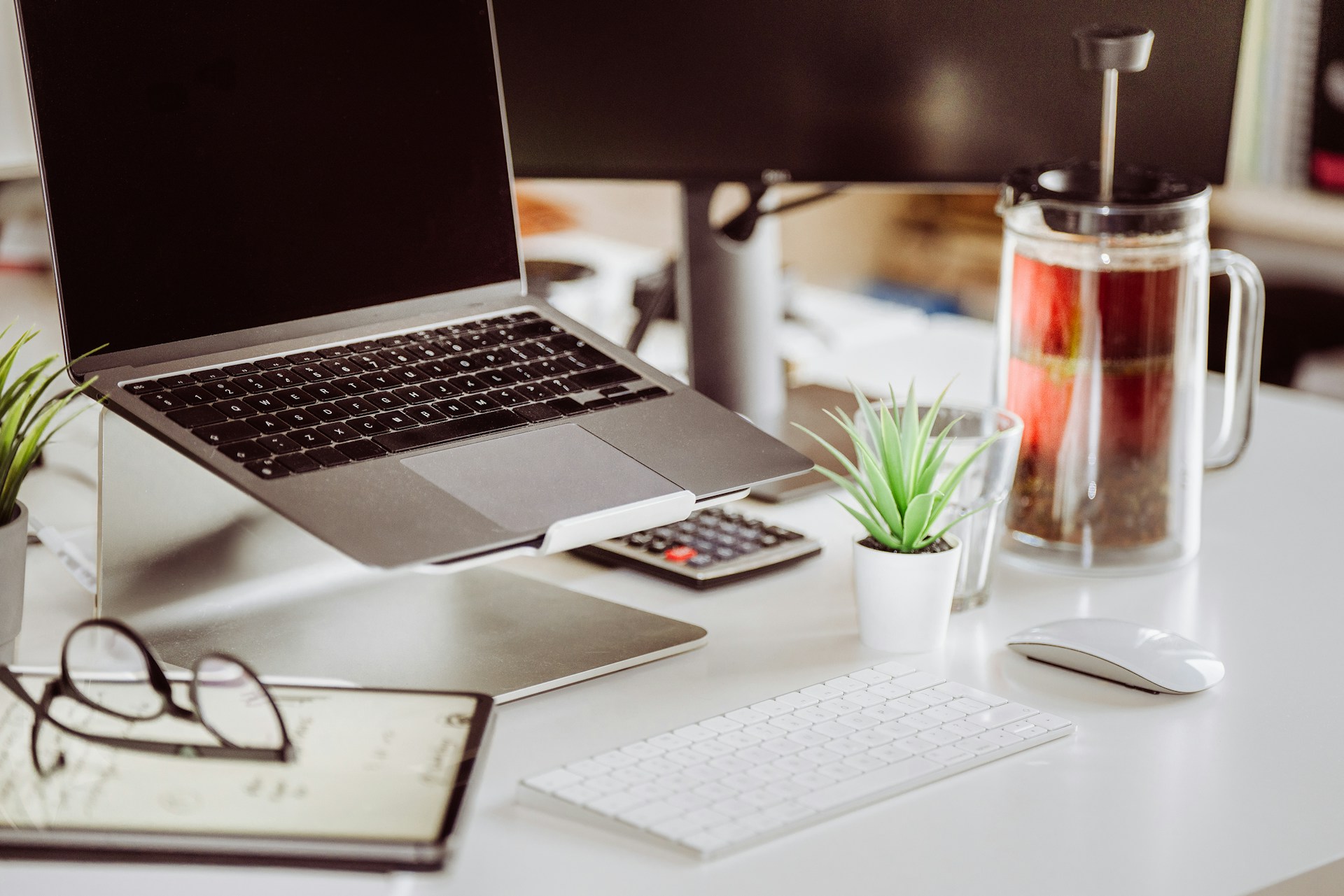 Modern office desk setup with a laptop on a stand, keyboard, mouse, and a French press coffee maker.