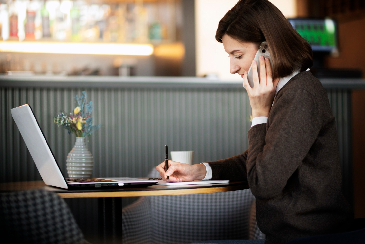 Professional woman talking on her smartphone and taking notes while working on a laptop in a cafe.
