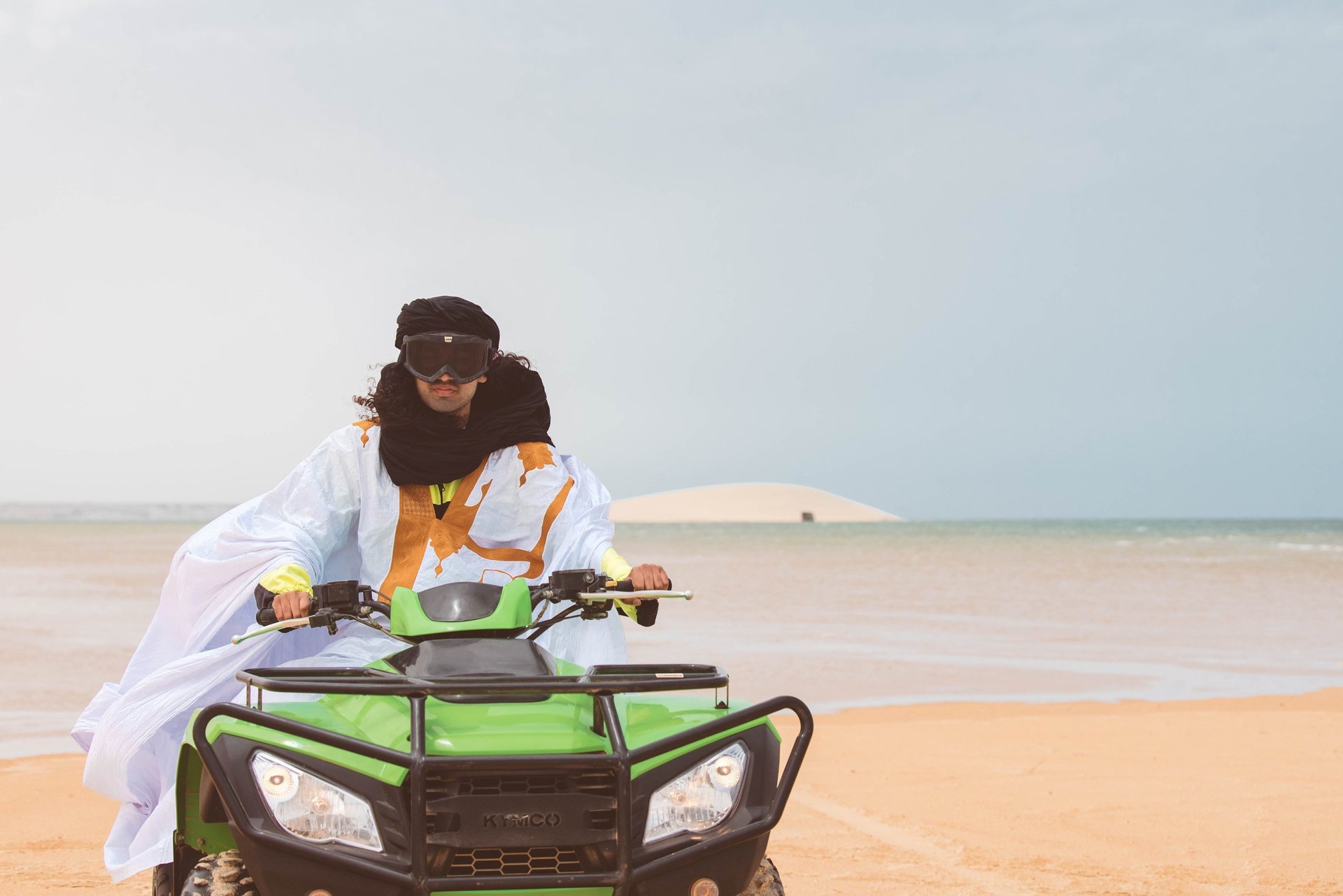 Man in traditional Sahraoui attire riding a lime green quad bike on the beach in Dakhla with the white dune in the background