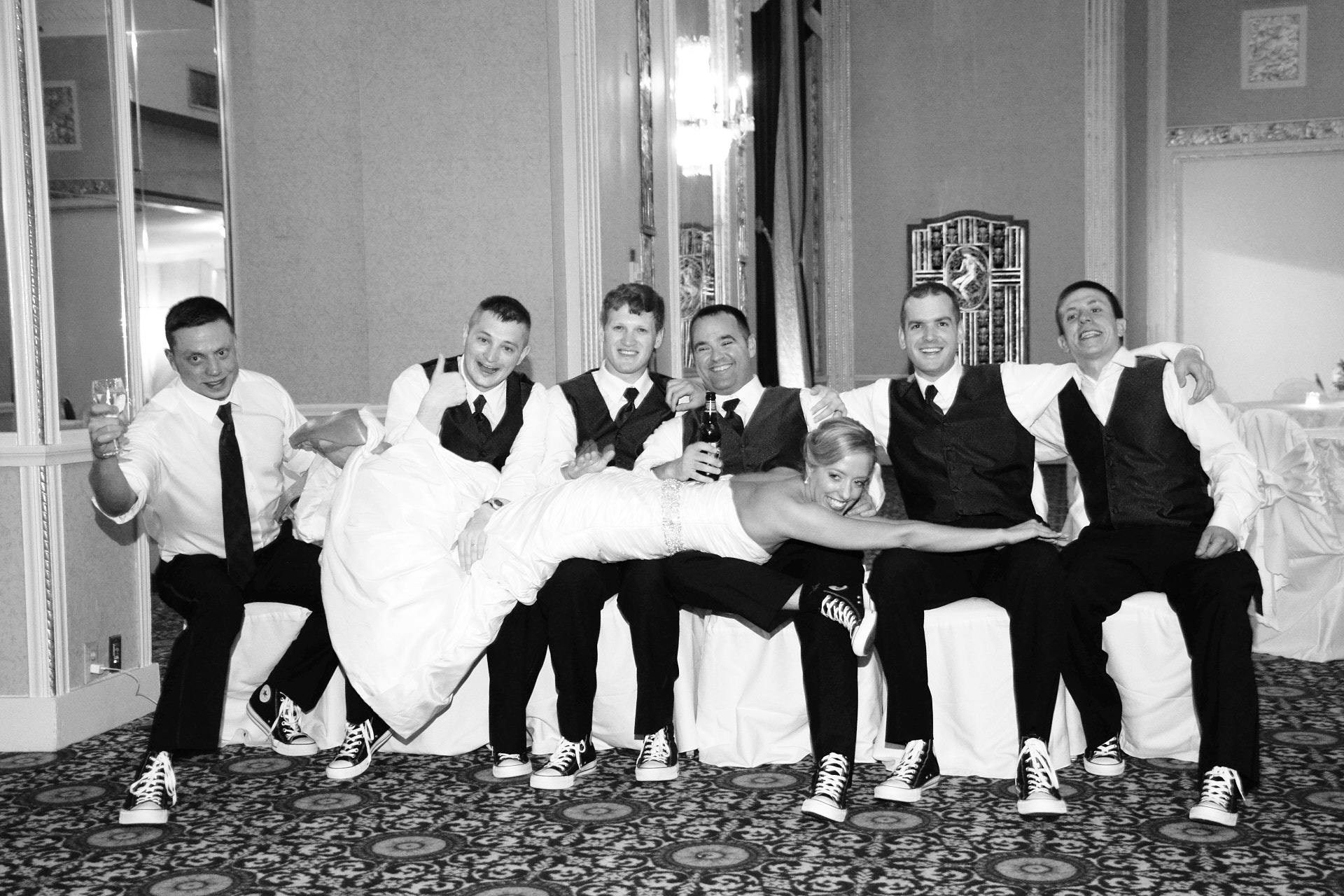 6 men in suits sitting on white chairs with a bride lying on them