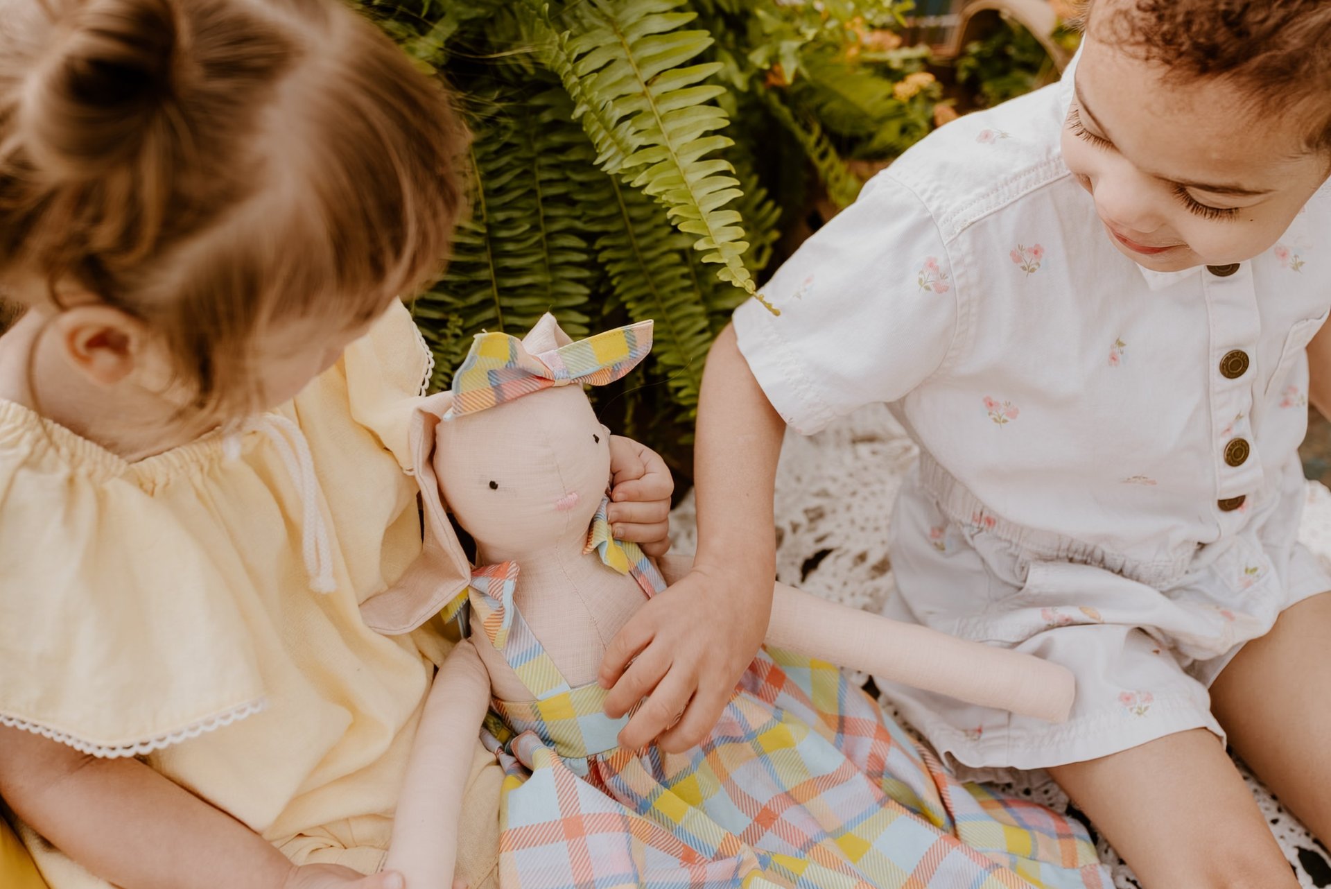Two young children playing with a soft linen bunny doll outdoors near a lush green fern.