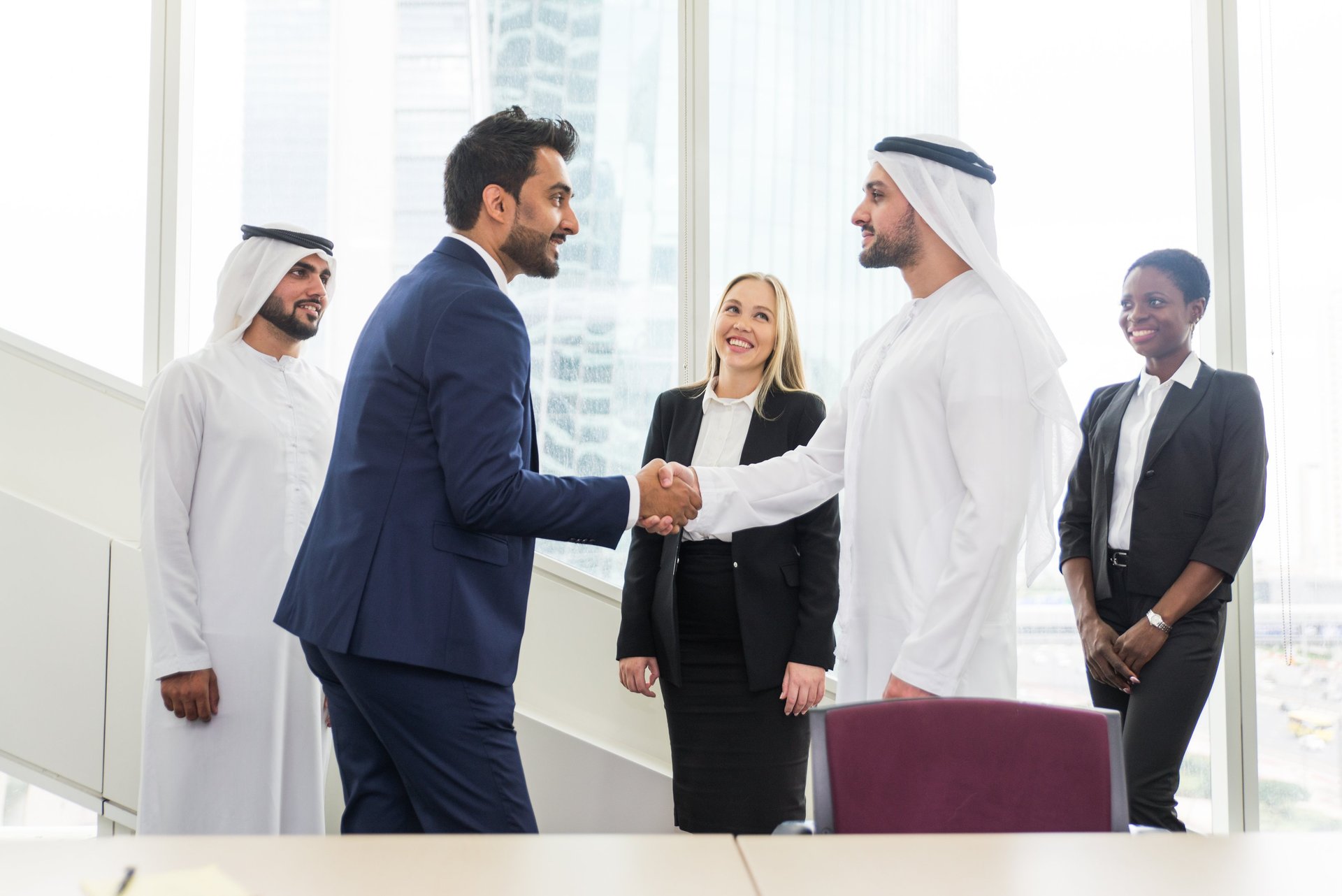 Businessman in a suit shakes hands with another in Emirati attire, while a businesswoman smiles