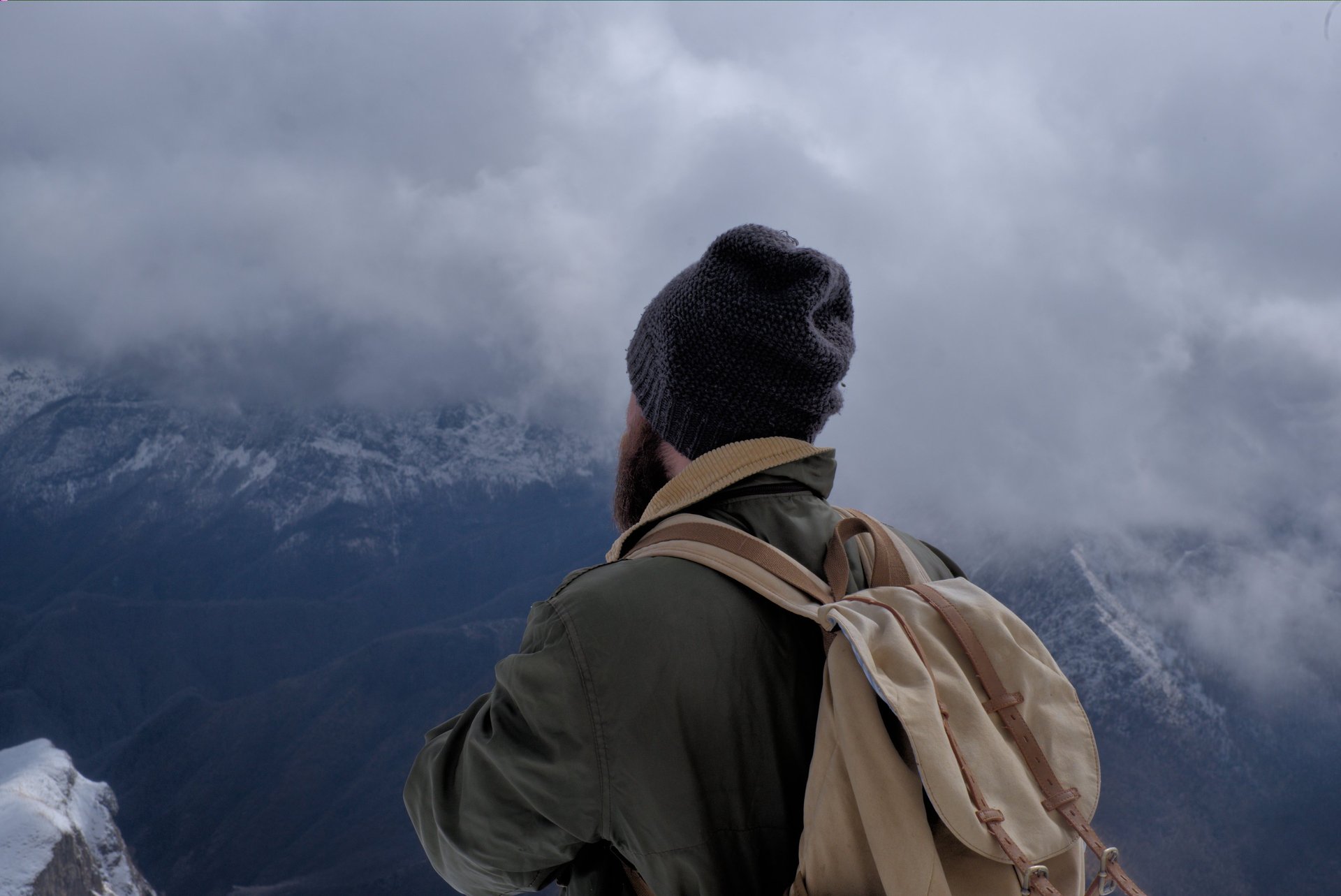A male hiker with a backpack and beanie overlooking a cloudy mountain range in winter.