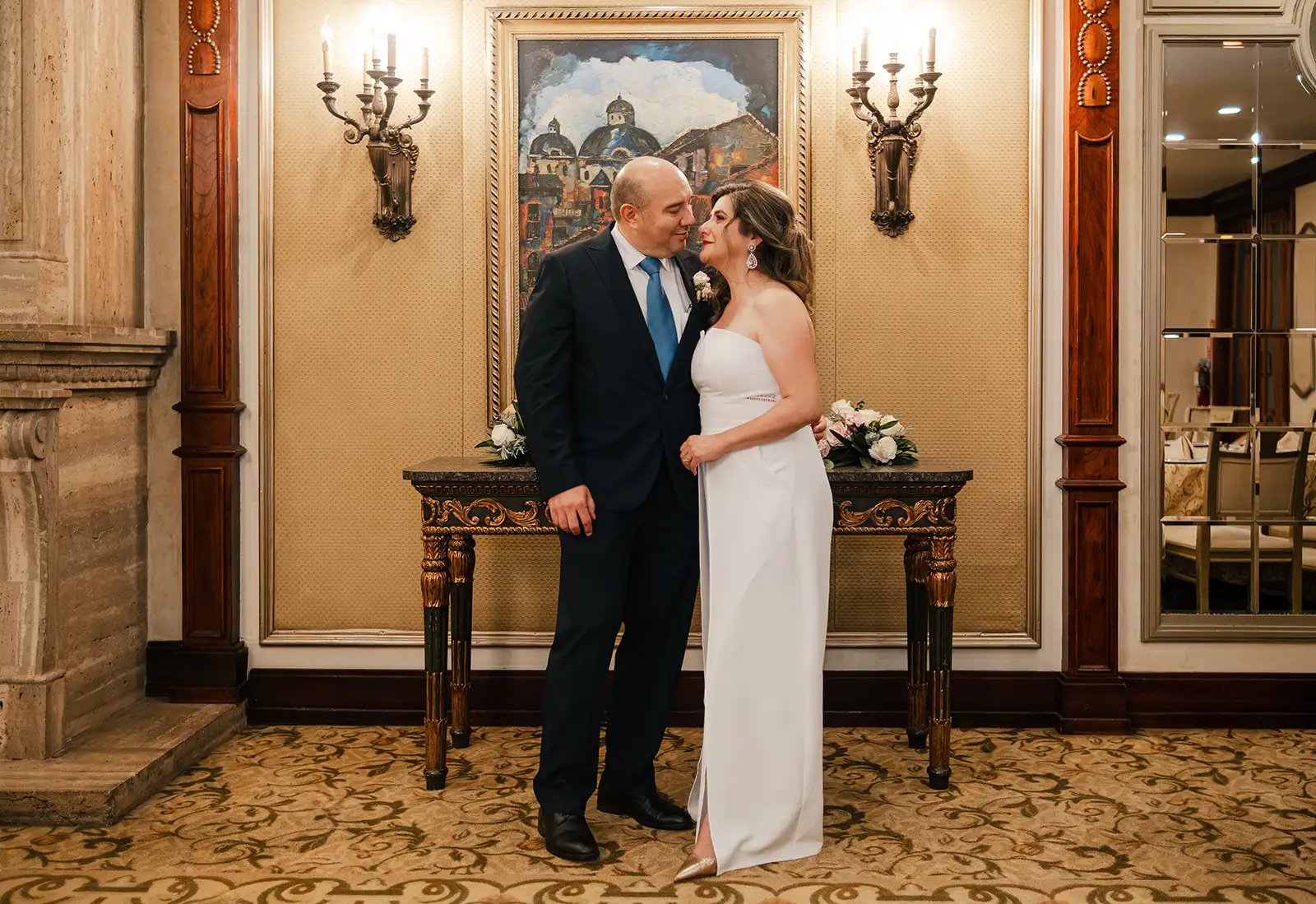 Hombre con traje junto novia con vestido blanco, viéndose mutuamente en el interior de una sala elegante con candelabros