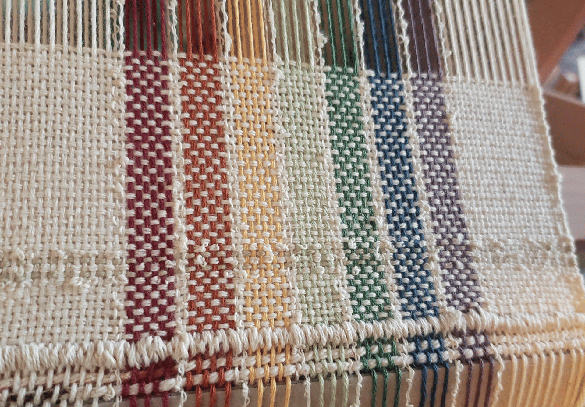 a loom with an unfinished hand-woven towel in muted rainbow pride flag colours