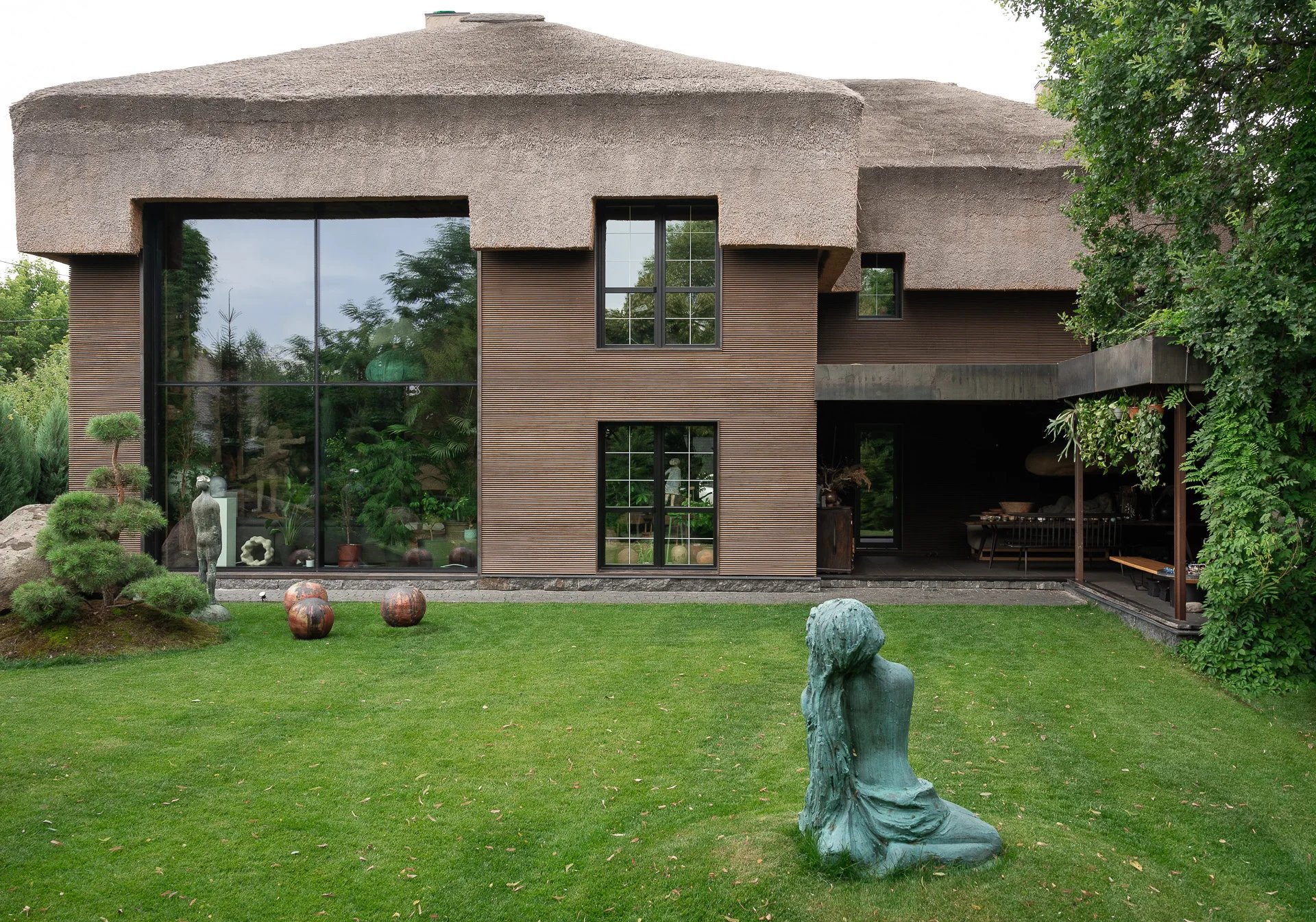 Nowoczesny dom jednorodzinny z dużymi przeszkleniami i strzechą, ogród i rzeźba na trawniku
