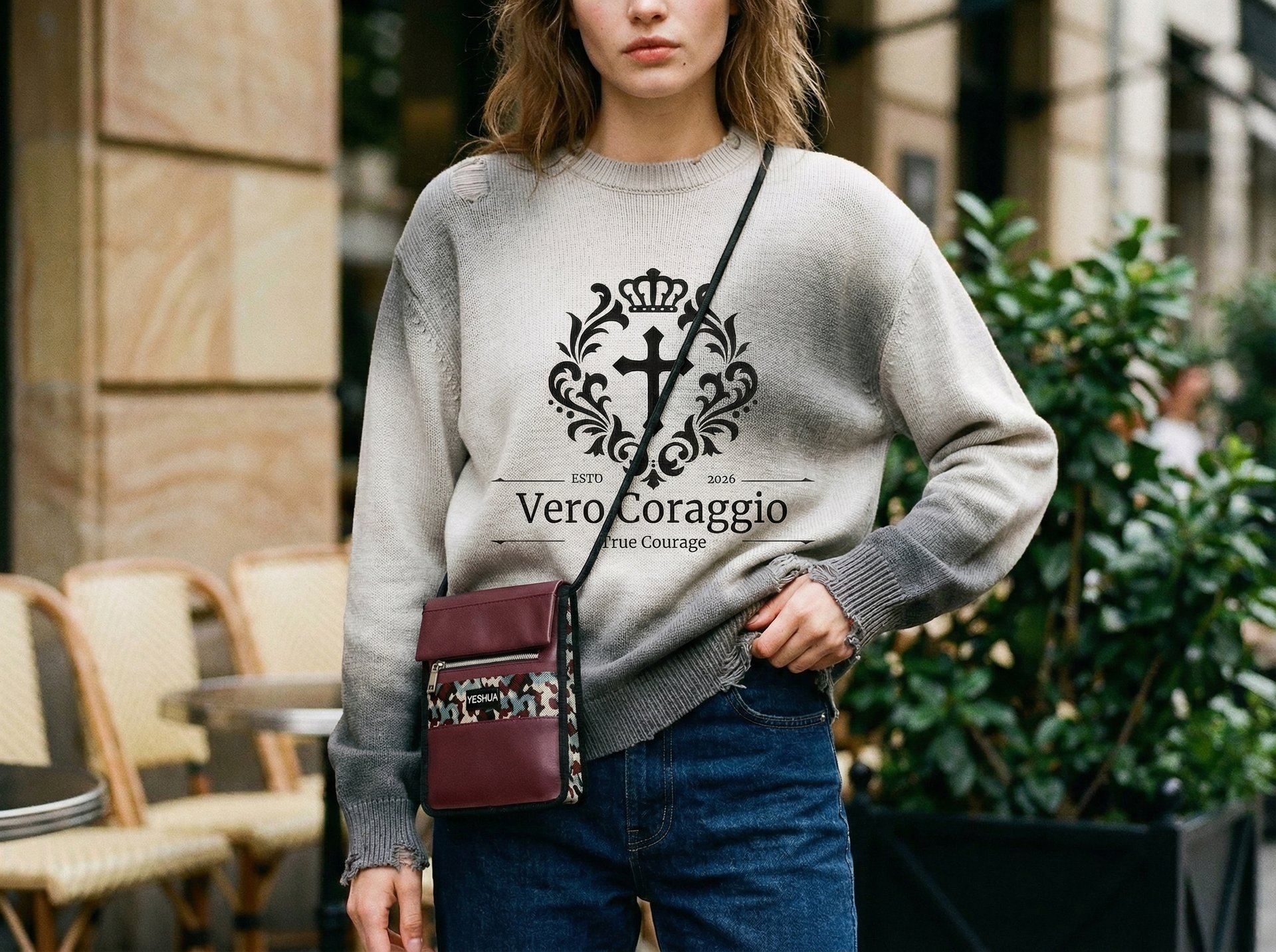 Woman wearing a grey Vero Coraggio graphic sweater with blue jeans and a burgundy crossbody bag.