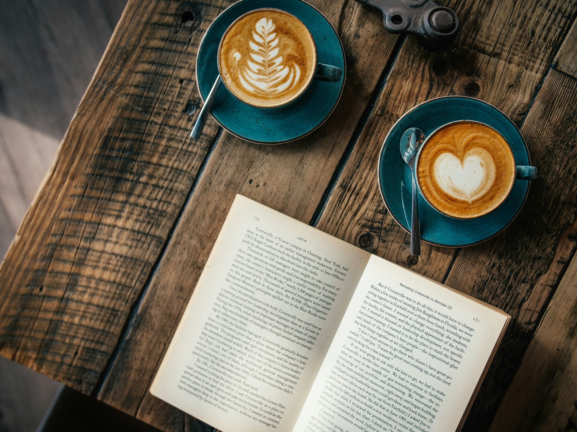 Two cups of latte art coffee sit on a rustic wooden table next to an open book in a cozy cafe.