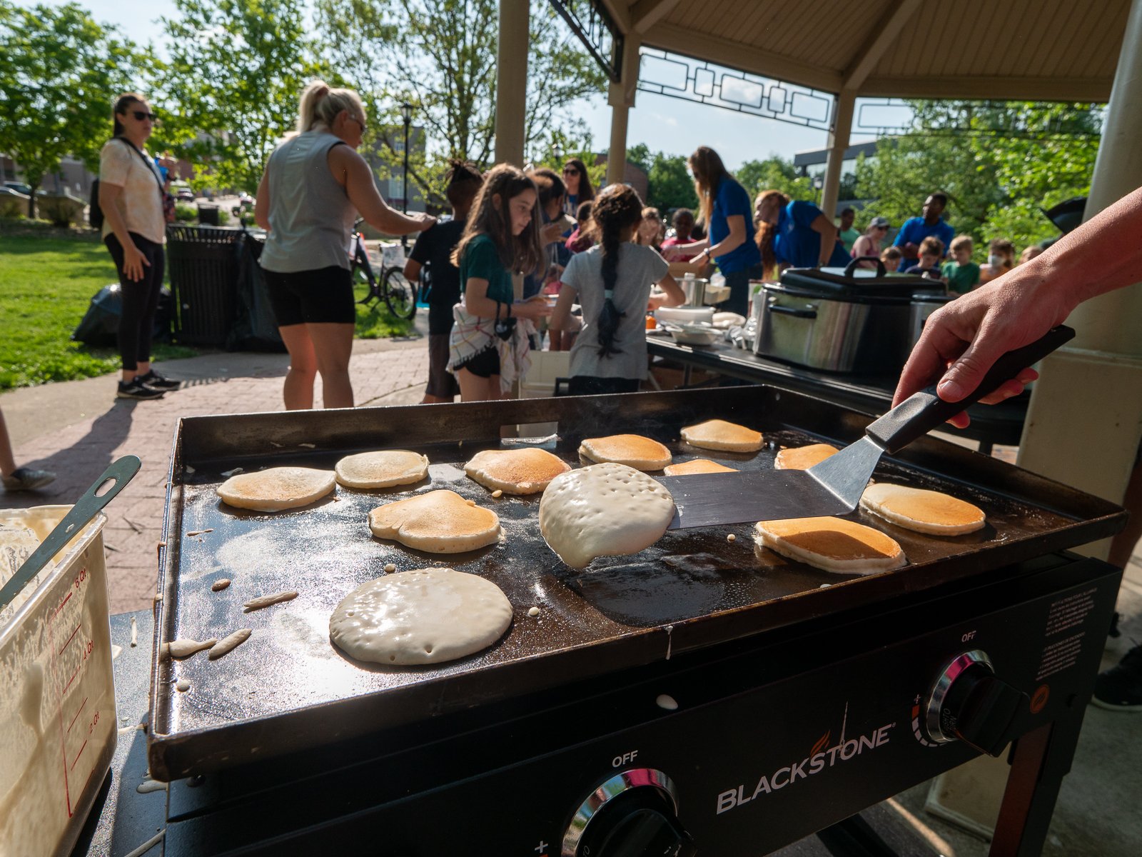 A hand flipping pancakes with a spatula, with people in the background