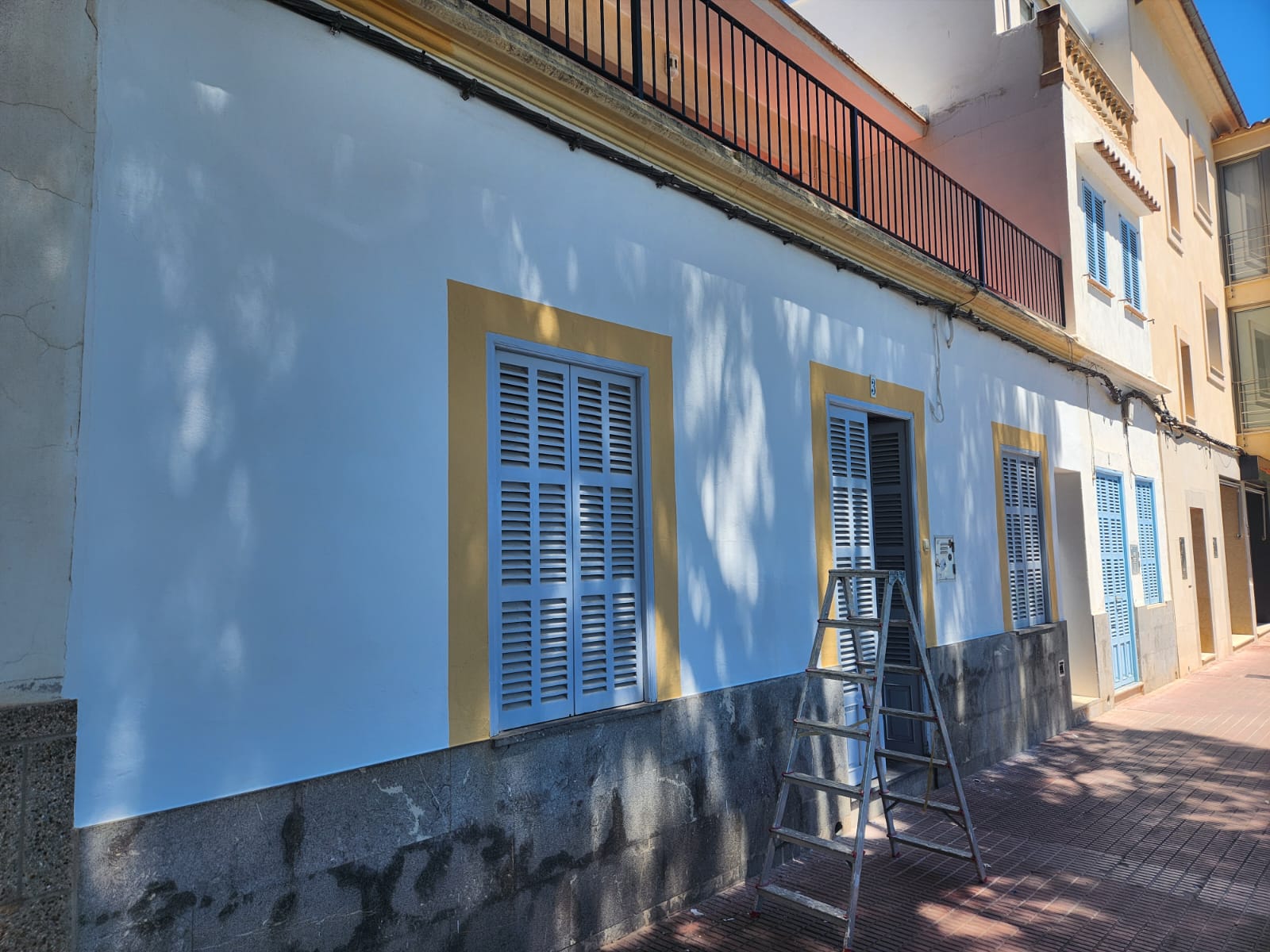 White stucco Mediterranean house with blue shutters and a metal ladder on the sidewalk.