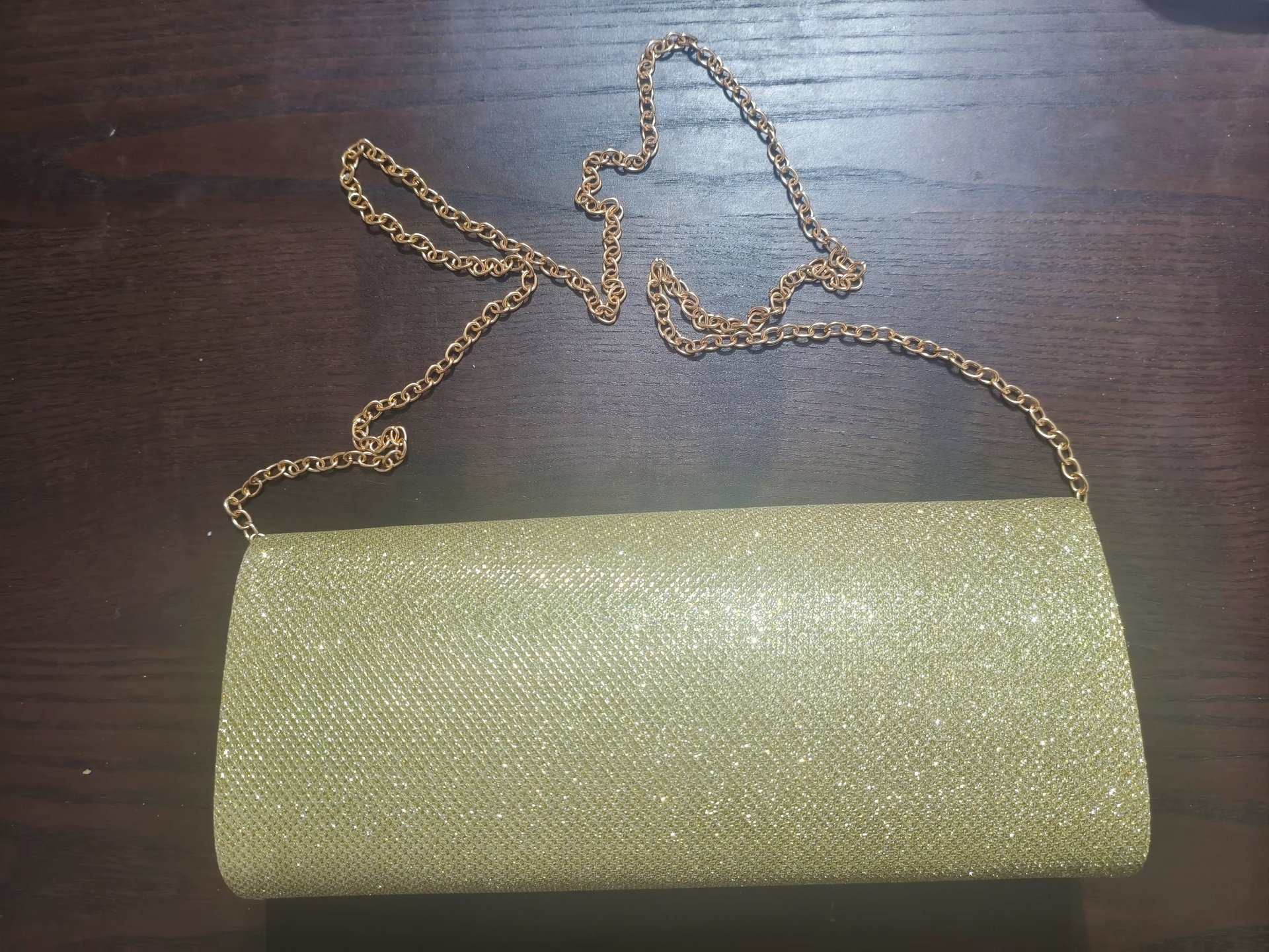 Mala clutch de festa dourada com glitter brilhante e alça de corrente metálica sobre fundo escuro.