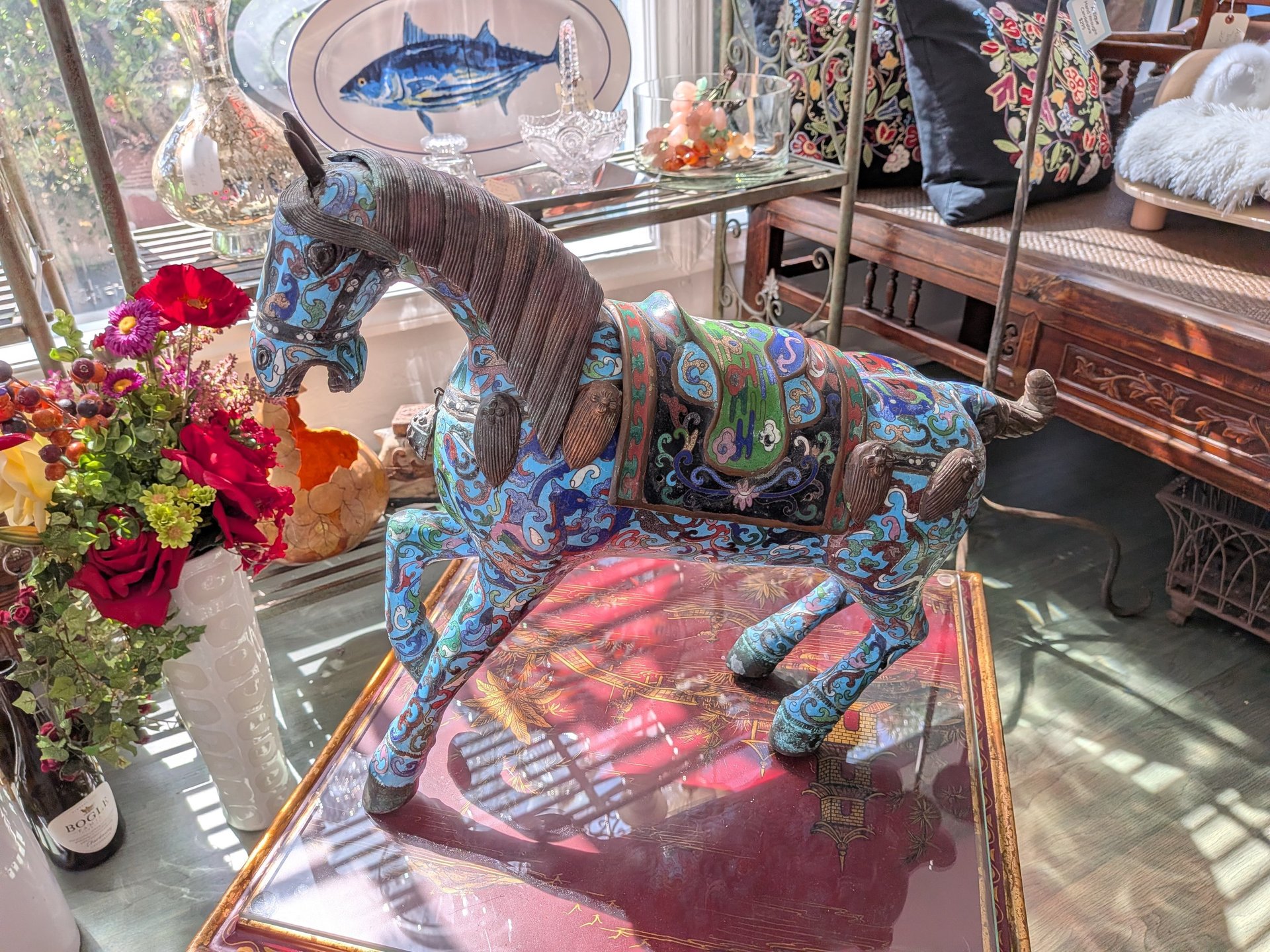 Ornate blue cloisonne horse statue with intricate floral patterns displayed in a sunlit antique shop.