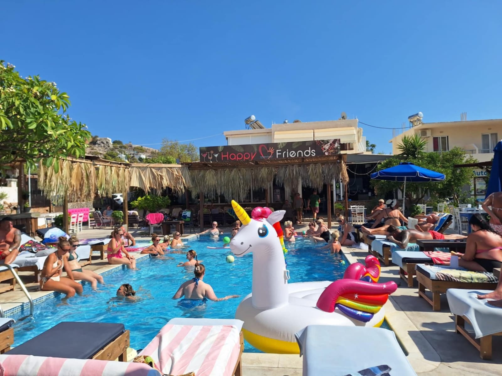 Crowded outdoor swimming pool at a resort with people relaxing and a large inflatable unicorn float.
