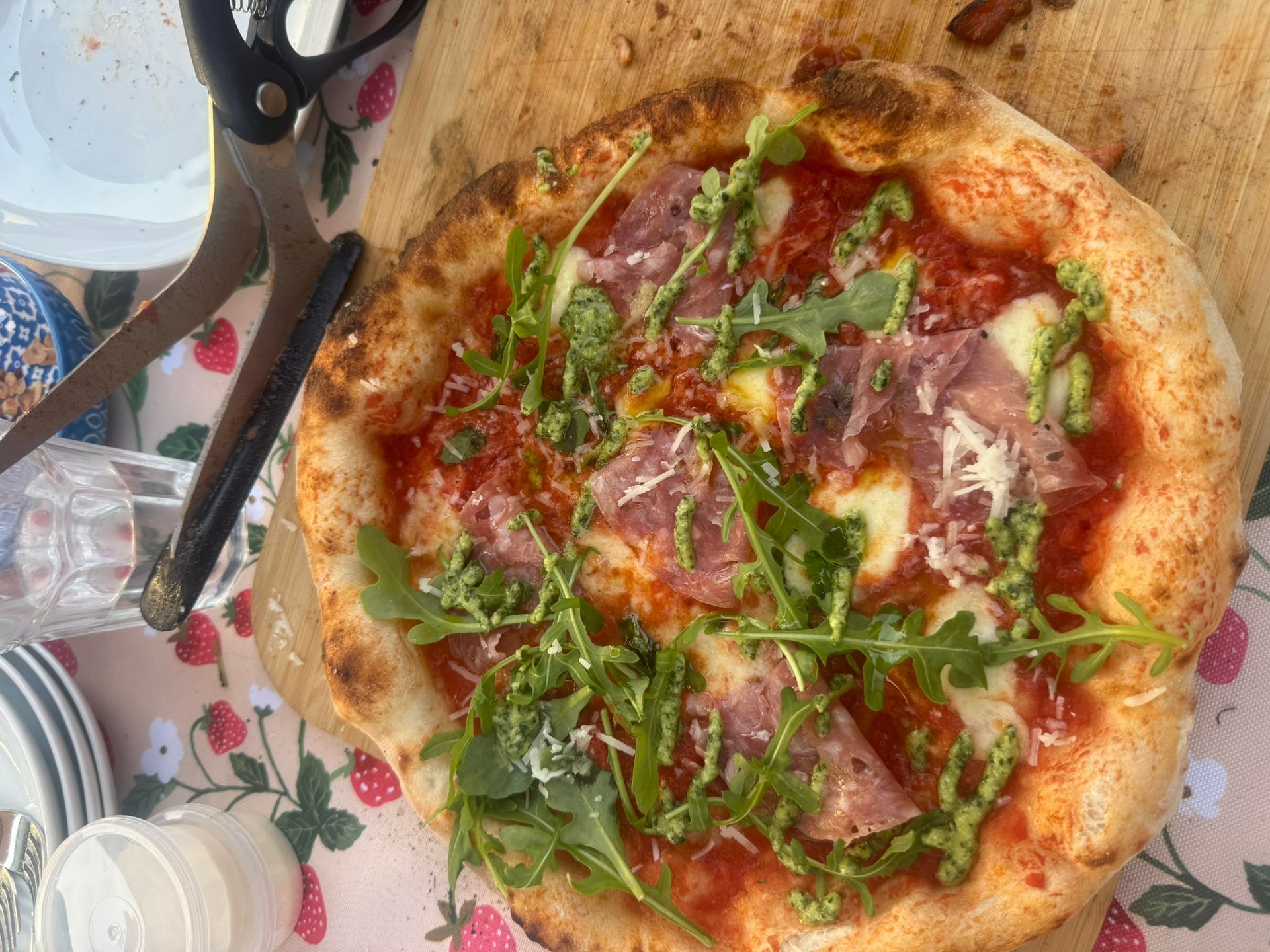 Homemade wood-fired pizza topped with prosciutto, fresh arugula, and pesto drizzle on a wooden board.