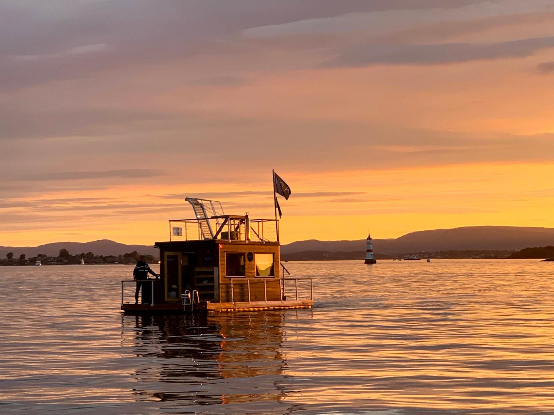 A small wooden houseboat floats on calm water during a golden sunset near a distant lighthouse and hills.