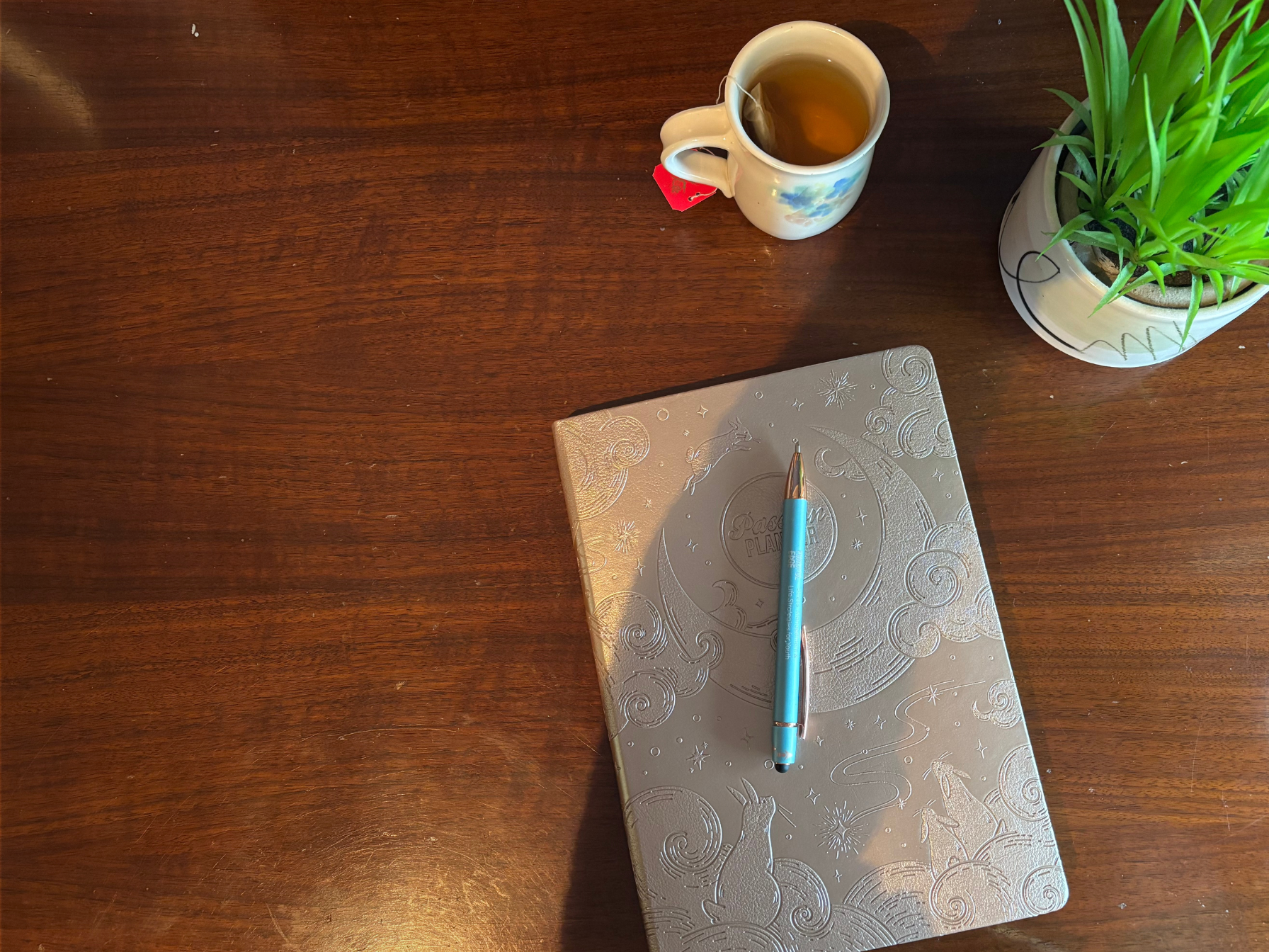 A silver embossed journal and blue pen on a wooden desk with a cup of tea and a potted green plant.