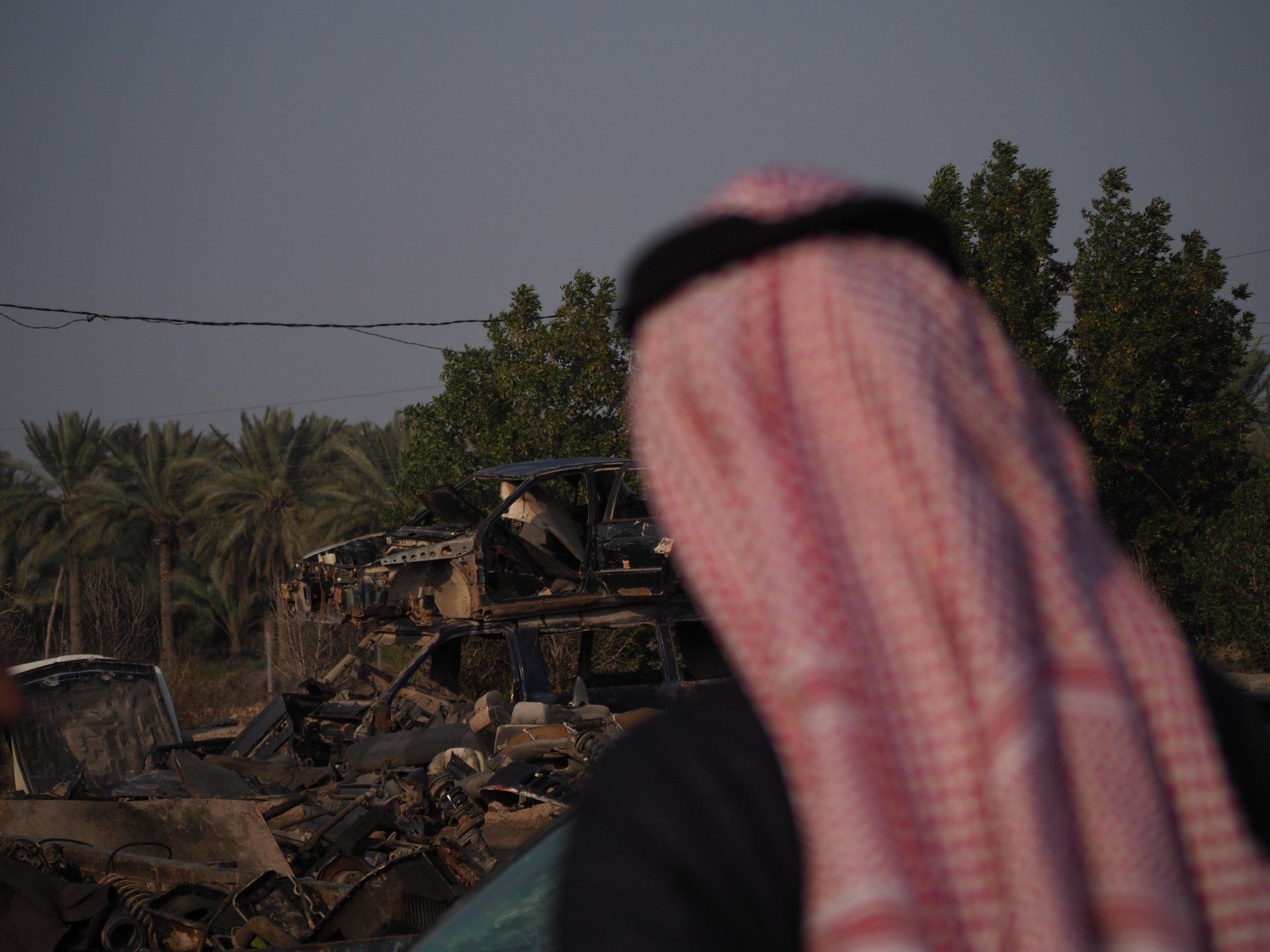 an arab man in a red and white scarf looking at a destroyed car in the by the palm trees in Iraq