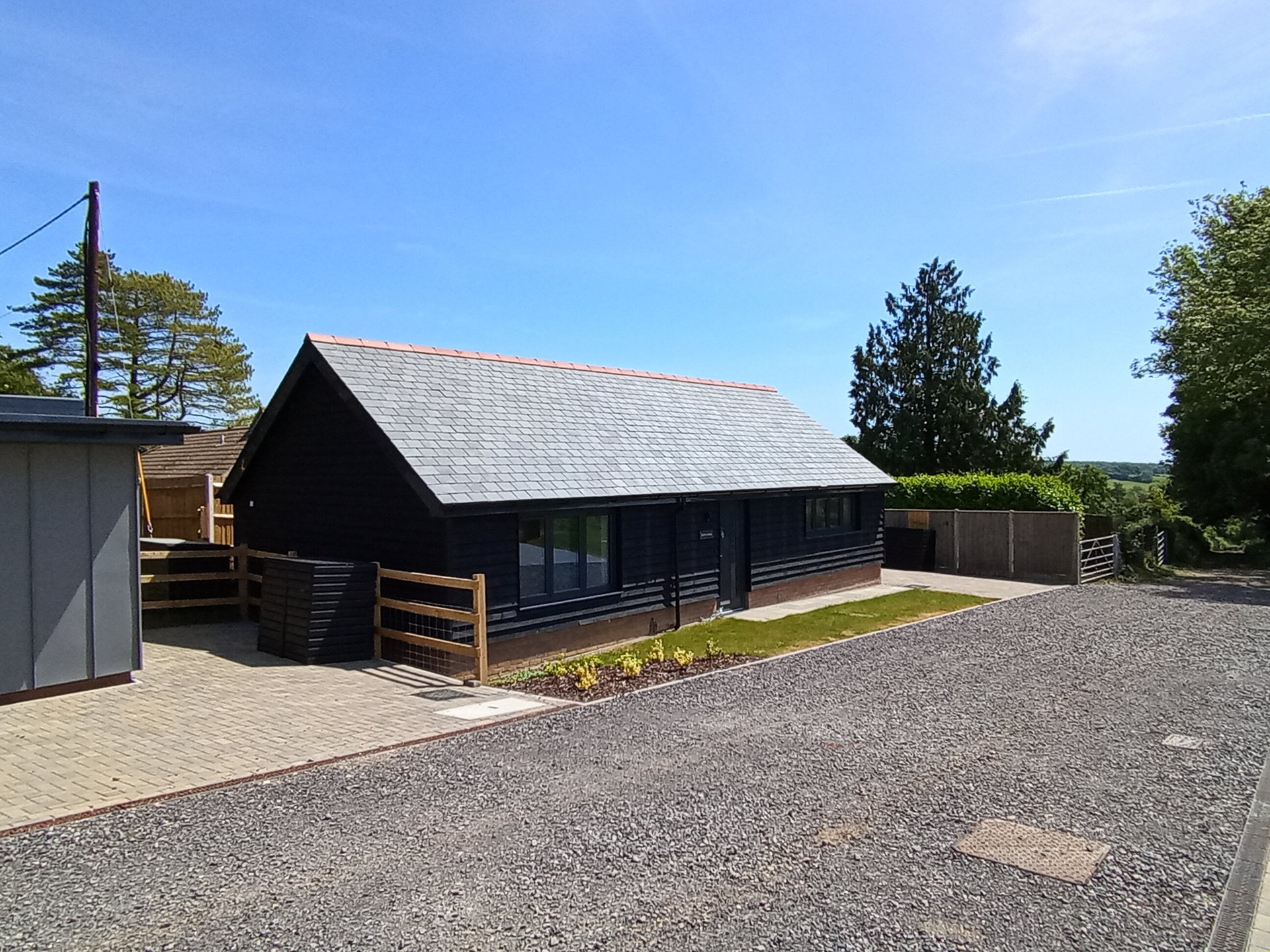 Modern black timber clad garden office with a grey slate roof next to a gravel driveway.
