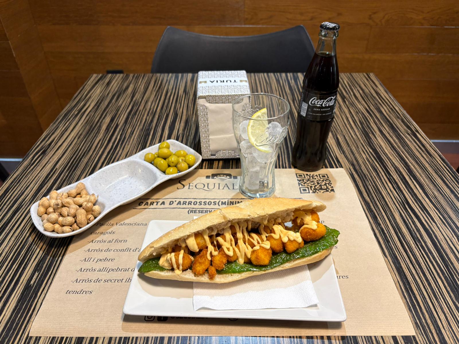 Spanish bocadillo sandwich with fried calamari and green sauce served with olives, peanuts, and a glass of ice.