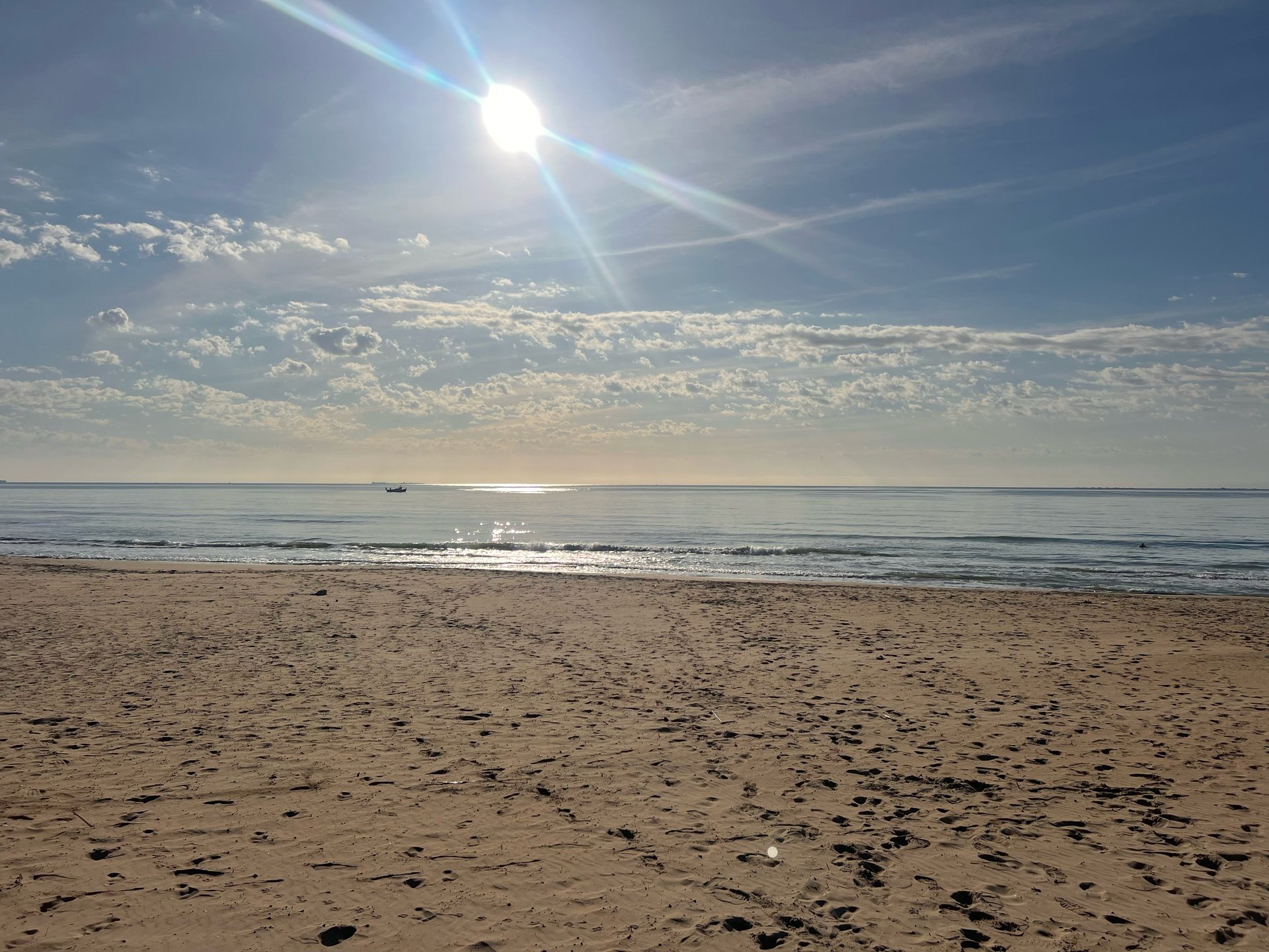 Peaceful morning by the Mediterranean Sea with a quiet beach, sparkling water, soft clouds and a small boat in the distance.