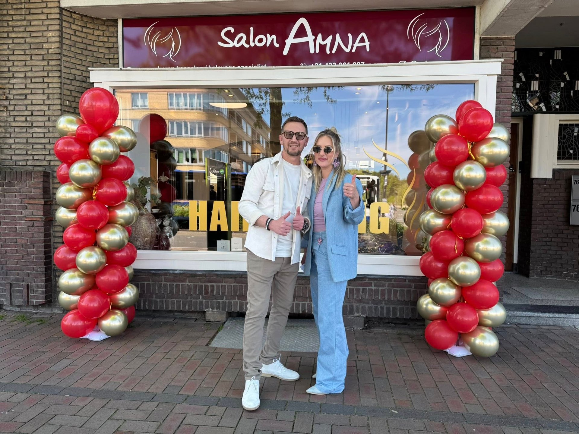 A couple posing in front of Salon Amna hair salon entrance decorated with red and gold balloon arches.