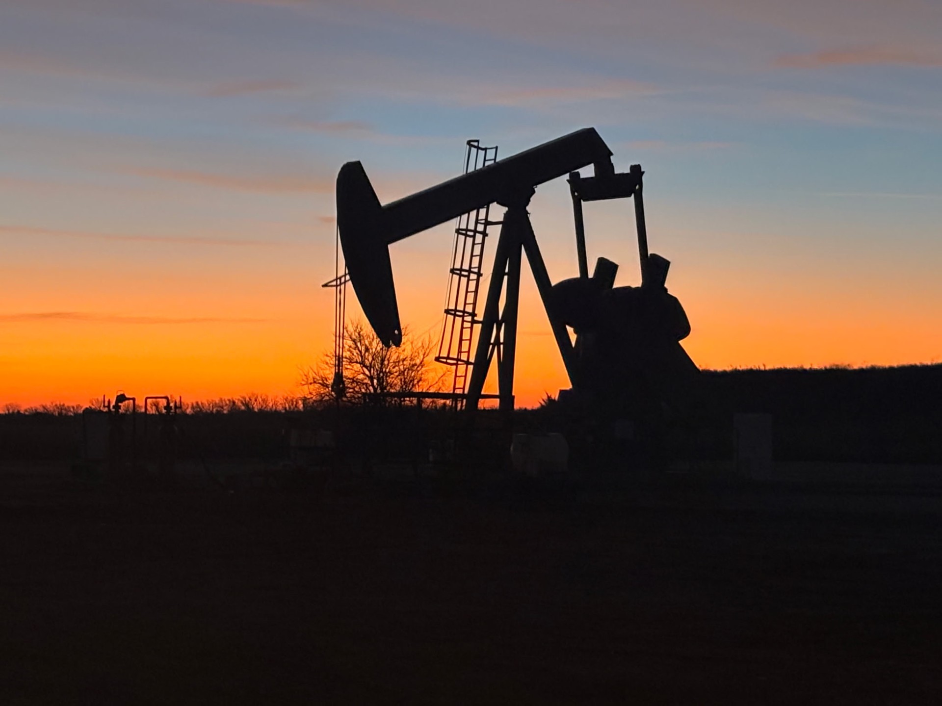 Silhouette of an oil pump jack extraction rig operating against a vibrant orange sunset horizon.